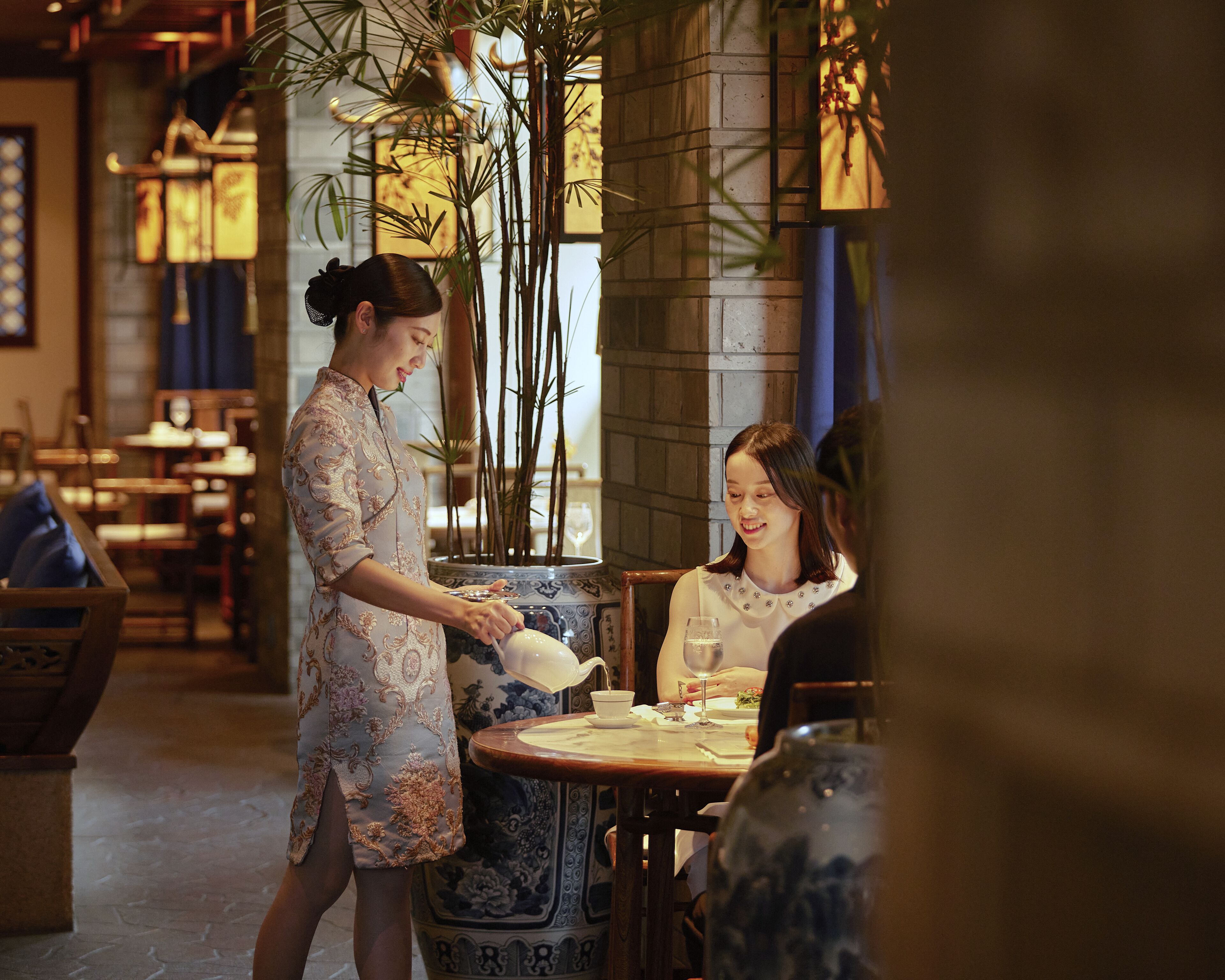 L’intérieur d’un restaurant, où l’on voit un serveur prendre la commande d’un client. On aperçoit des tables avec des chaises, un grand vase et une plante en pot.