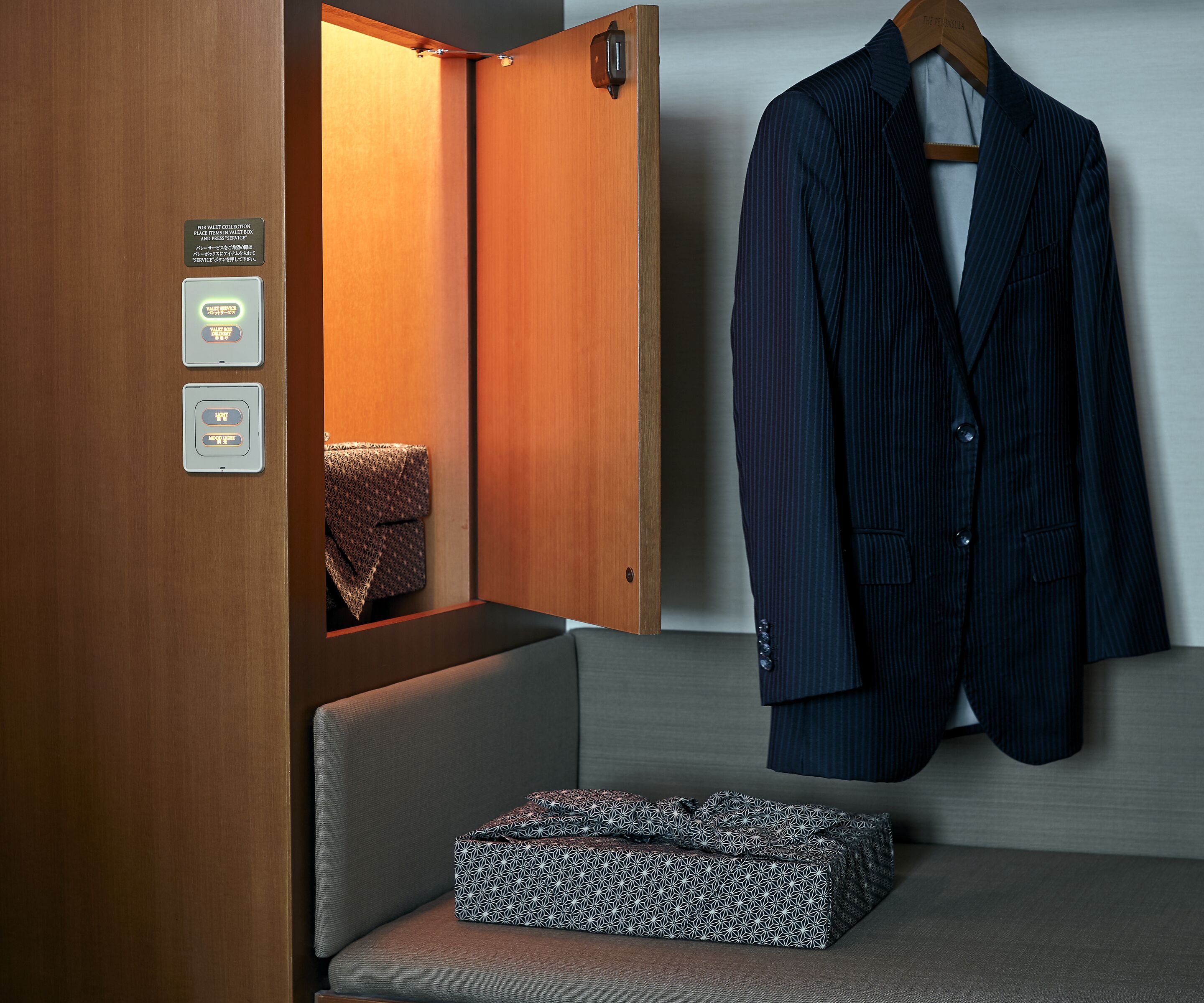 A hotel room with a wooden wardrobe, a hanging coat, and a patterned box on a cushioned bench.