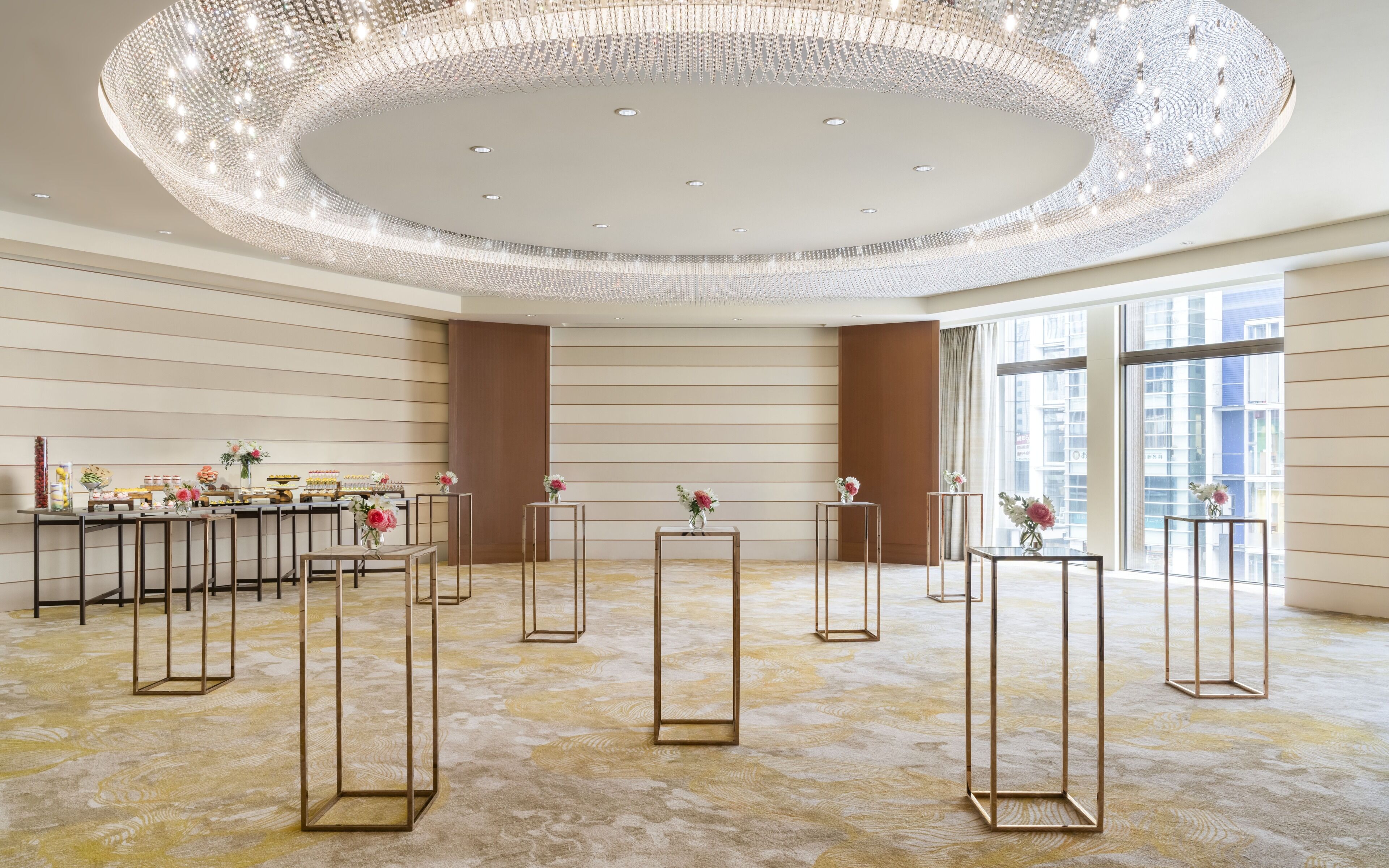 A spacious hotel lobby with a large chandelier, multiple tables with vases, and a view of the city through large windows.