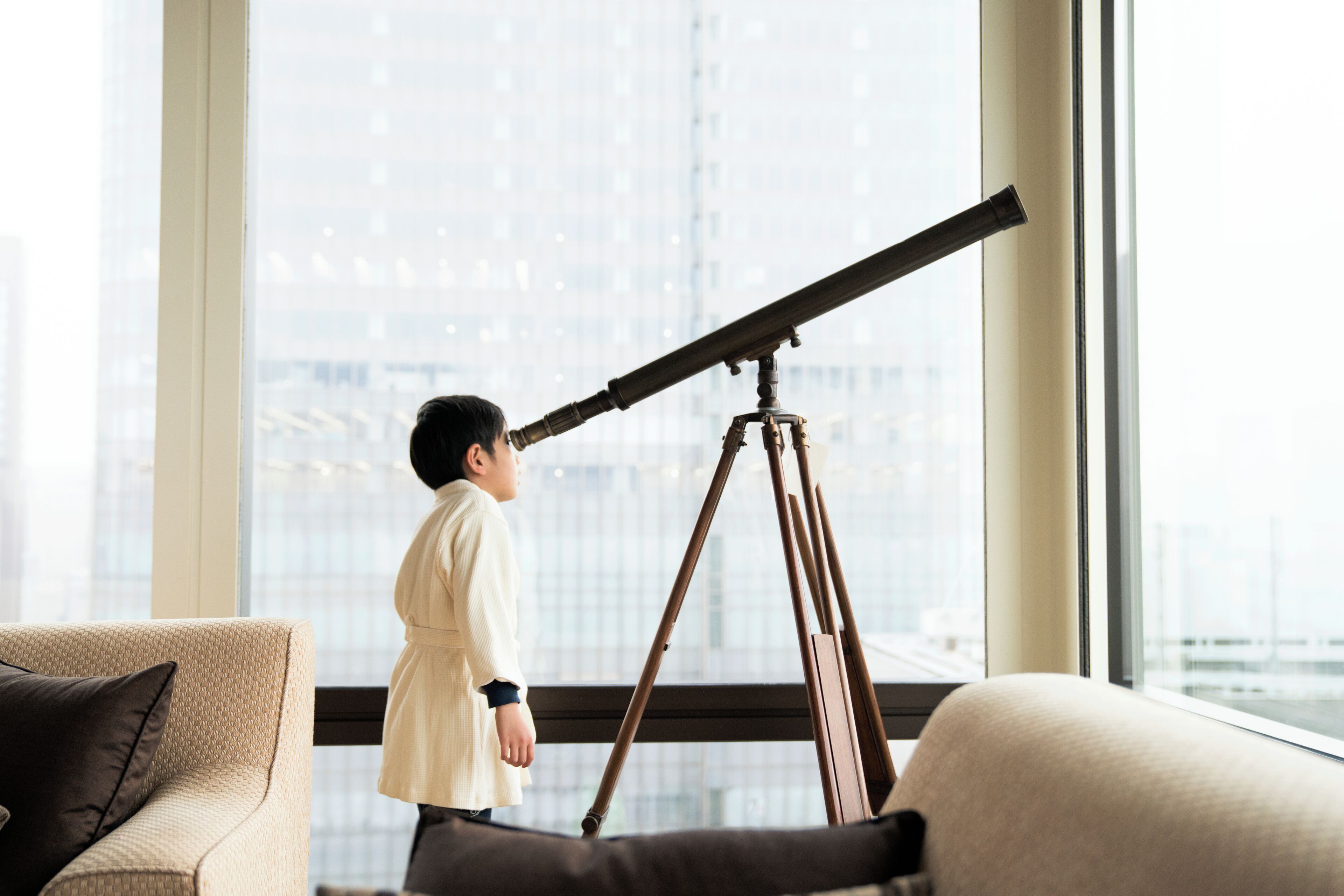 Una persona usando un telescopio en una habitación con ventanas amplias, un sofá y vista a la ciudad.