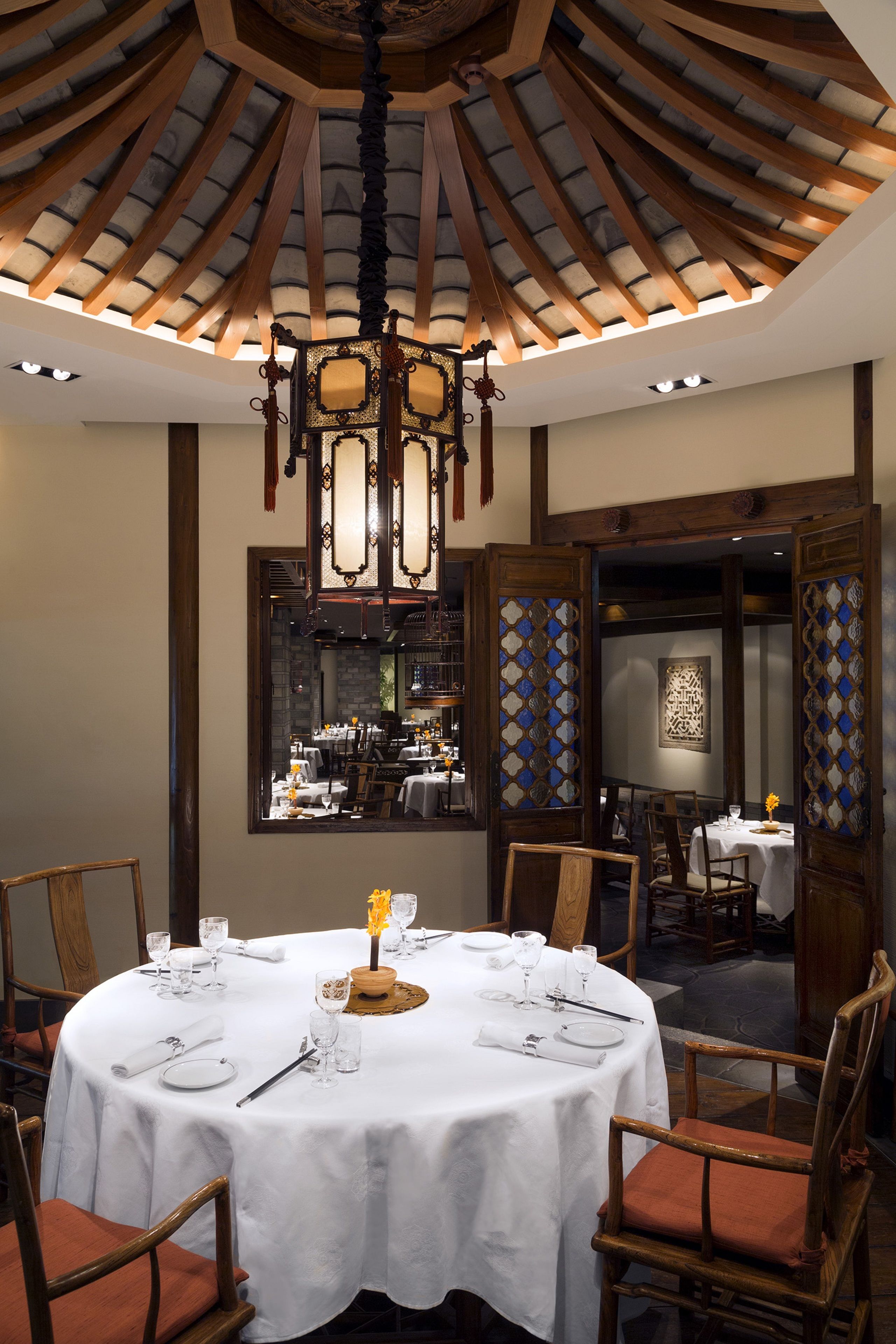 A dining area with a round table set for four, surrounded by wooden chairs. A chandelier hangs from the ceiling, and a decorative screen with a geometric pattern is visible in the background.