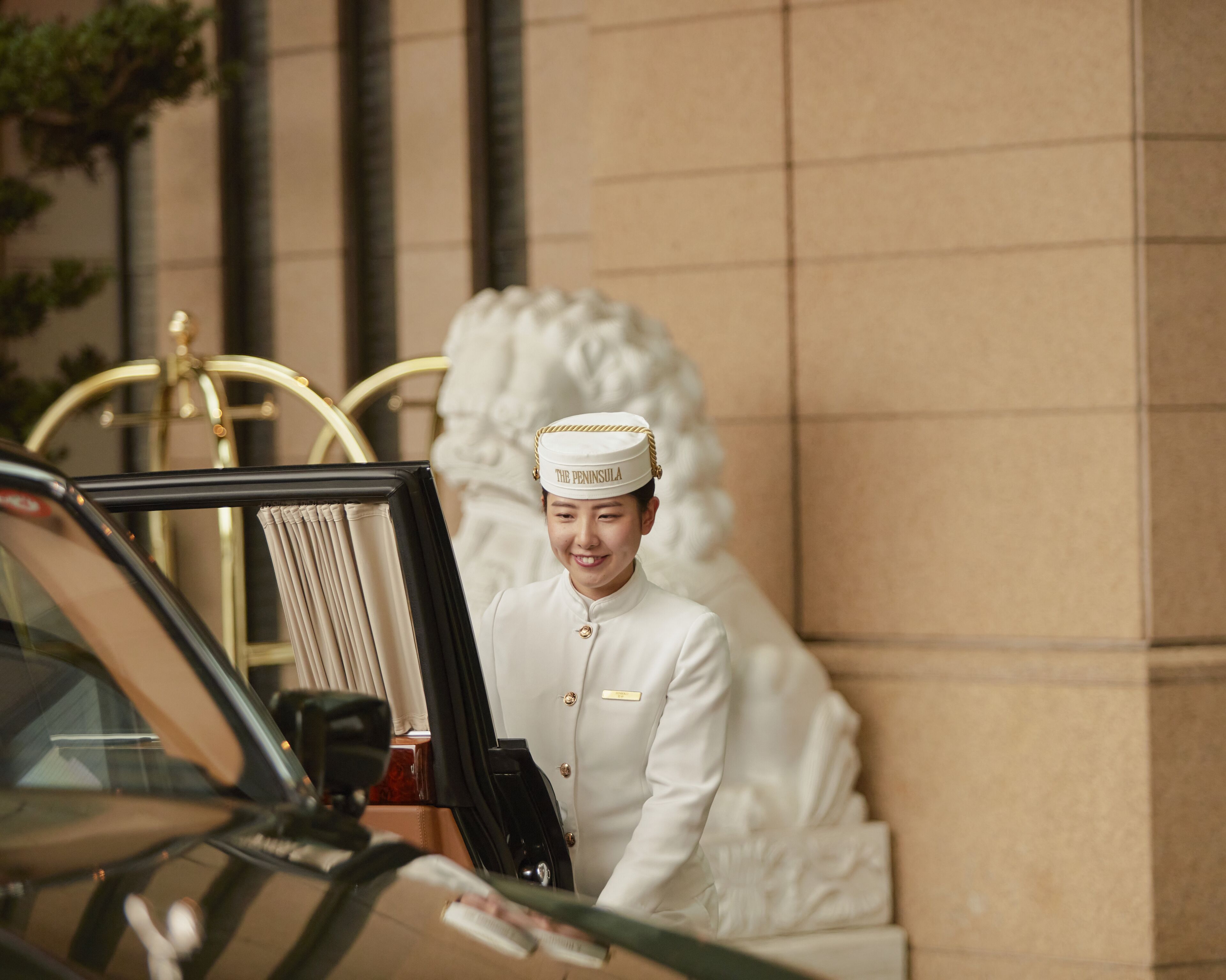 Un membre du personnel d’un hôtel, en uniforme, se tient à côté d’une voiture dont la porte porte une plaque avec un nom.