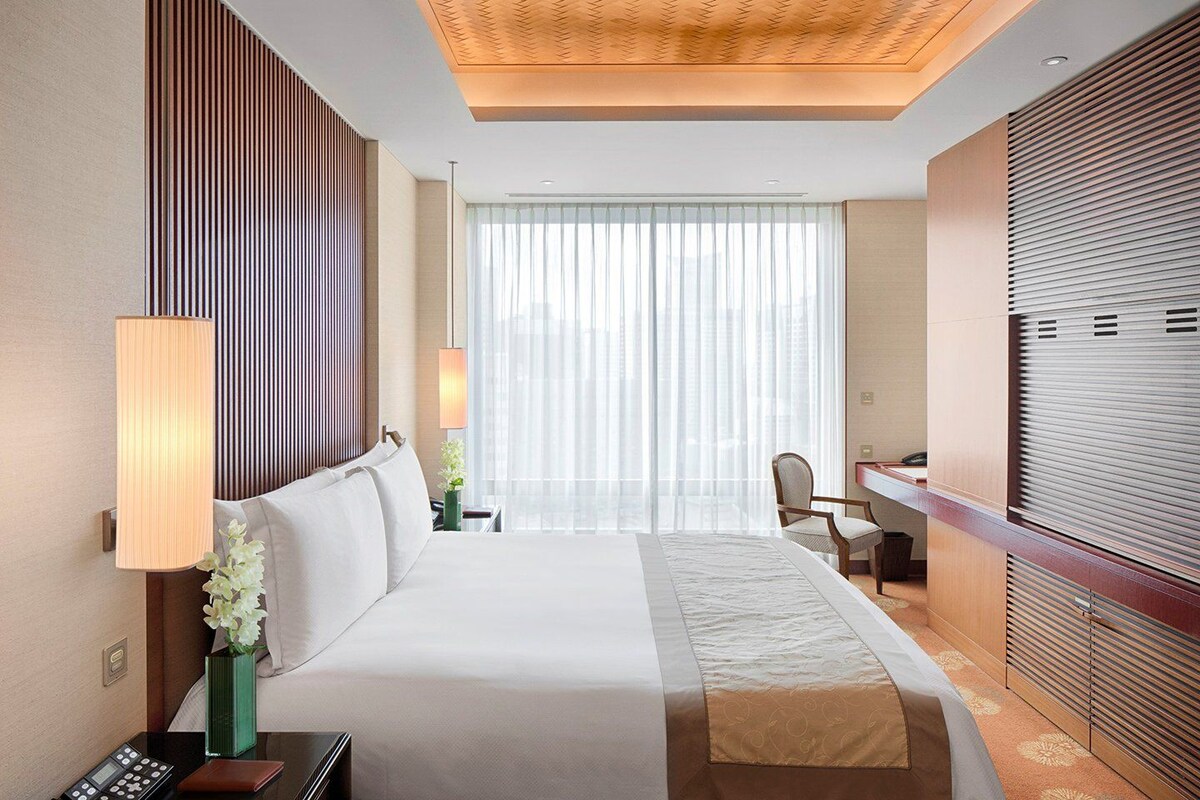 Une chambre d’hôtel comprenant un lit, une table de chevet, une lampe, une chaise, un bureau et une grande fenêtre avec des stores.