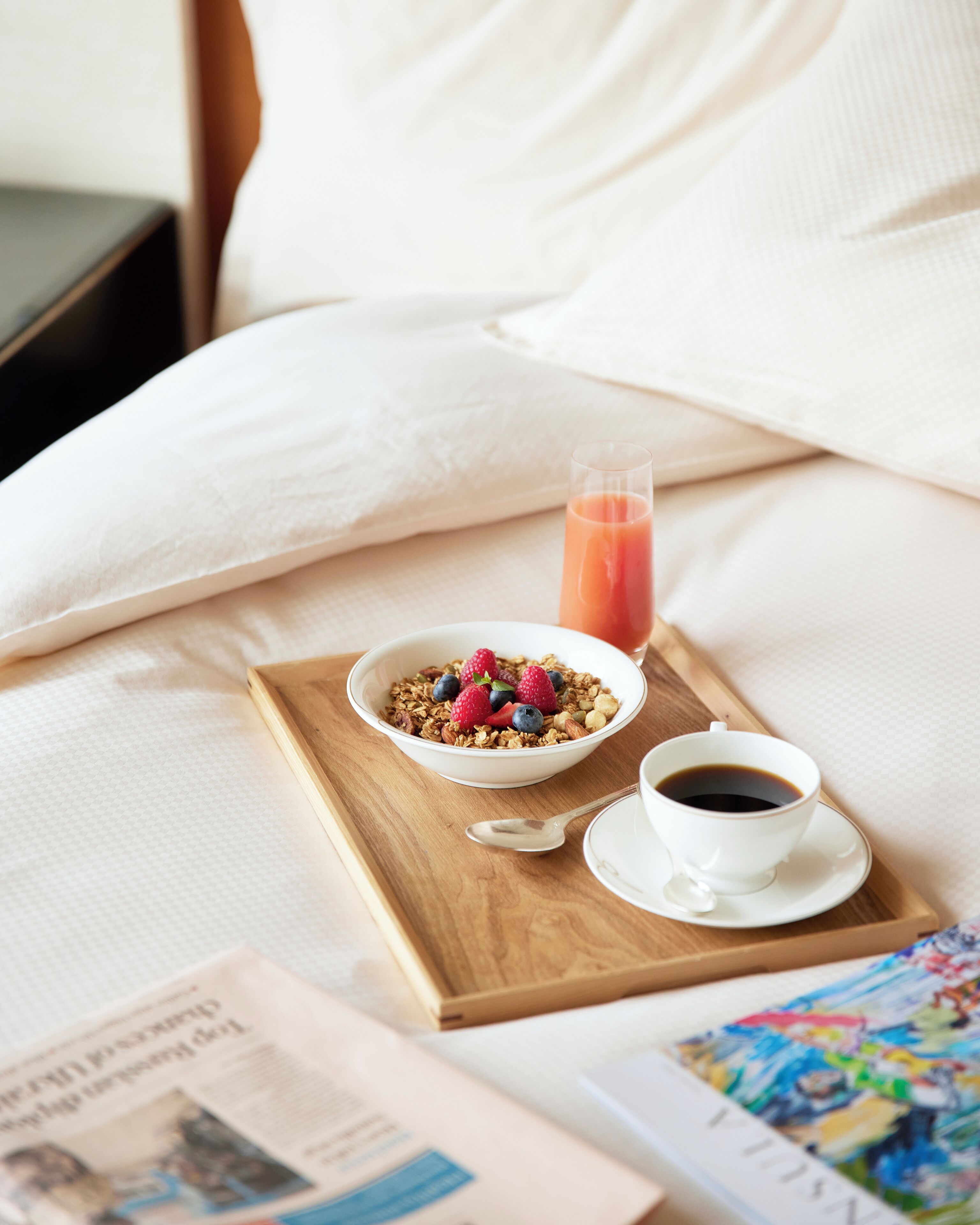 A breakfast tray with a bowl of granola and berries, a glass of orange juice, and a cup of coffee on a bed with white bedding.