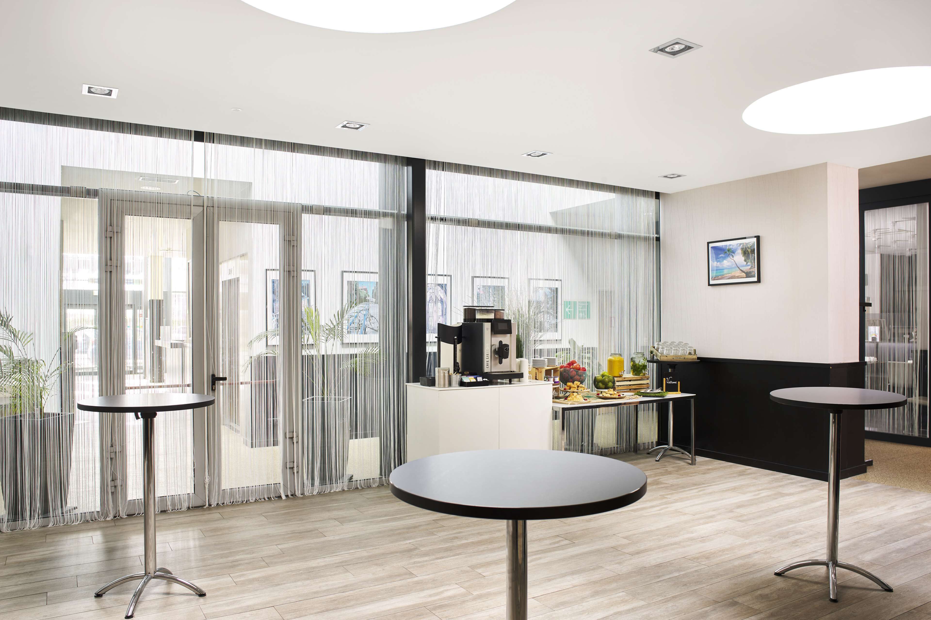 Un moderno vestíbulo de hotel con amplios ventanales, una estación de café y dos mesas redondas.