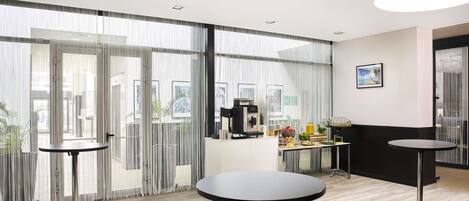 Un moderno vestíbulo de hotel con amplios ventanales, una estación de café y dos mesas redondas.