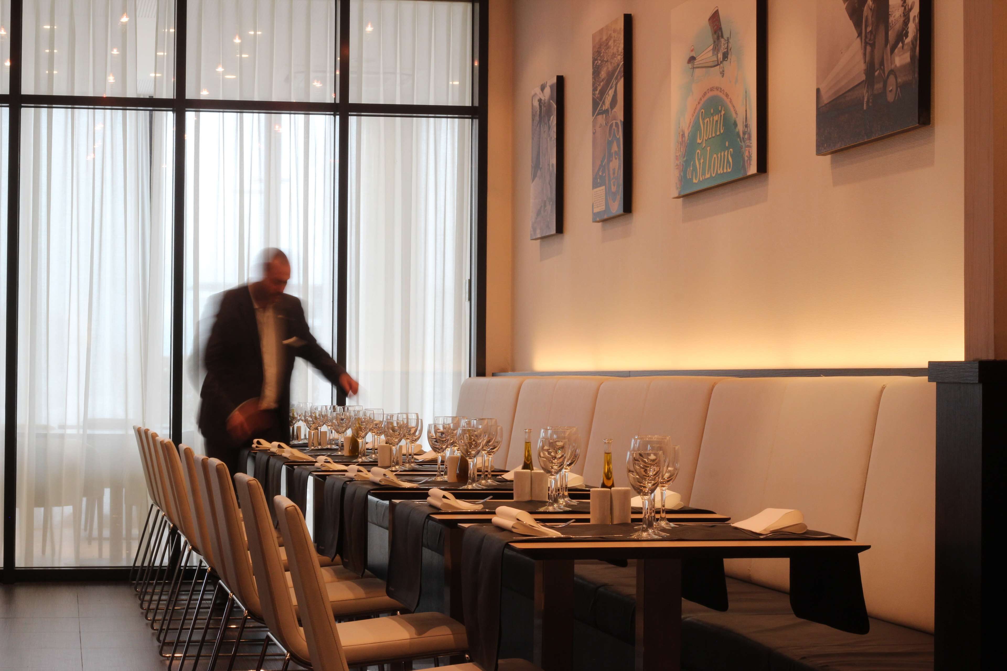 Un restaurante con una larga mesa lista para una comida, una zona de bar y un amplio ventanal con cortinas.