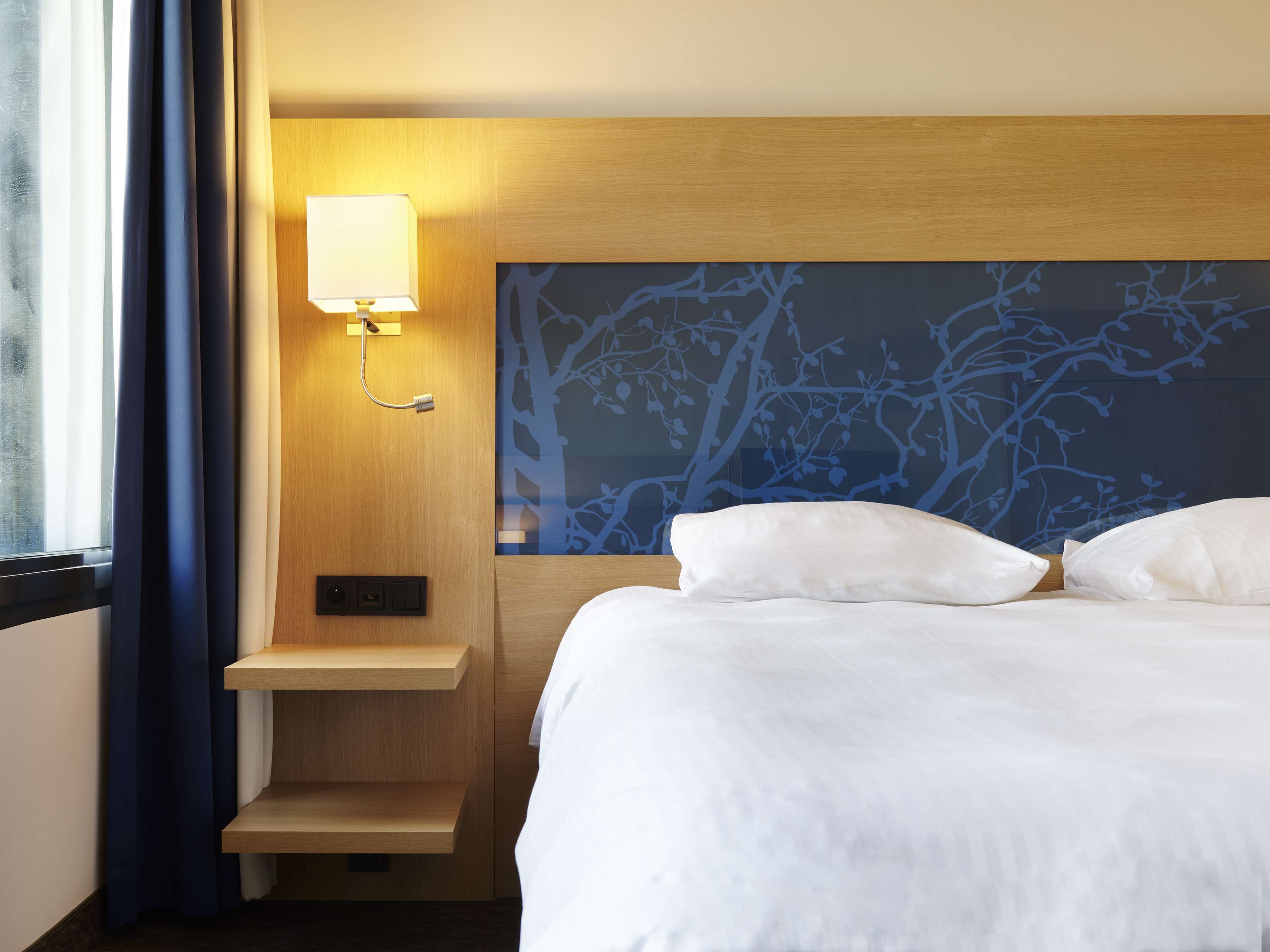 Ein Hotelzimmer mit Bett, Nachttisch, Wandlampe und einem Fenster mit Vorhängen.