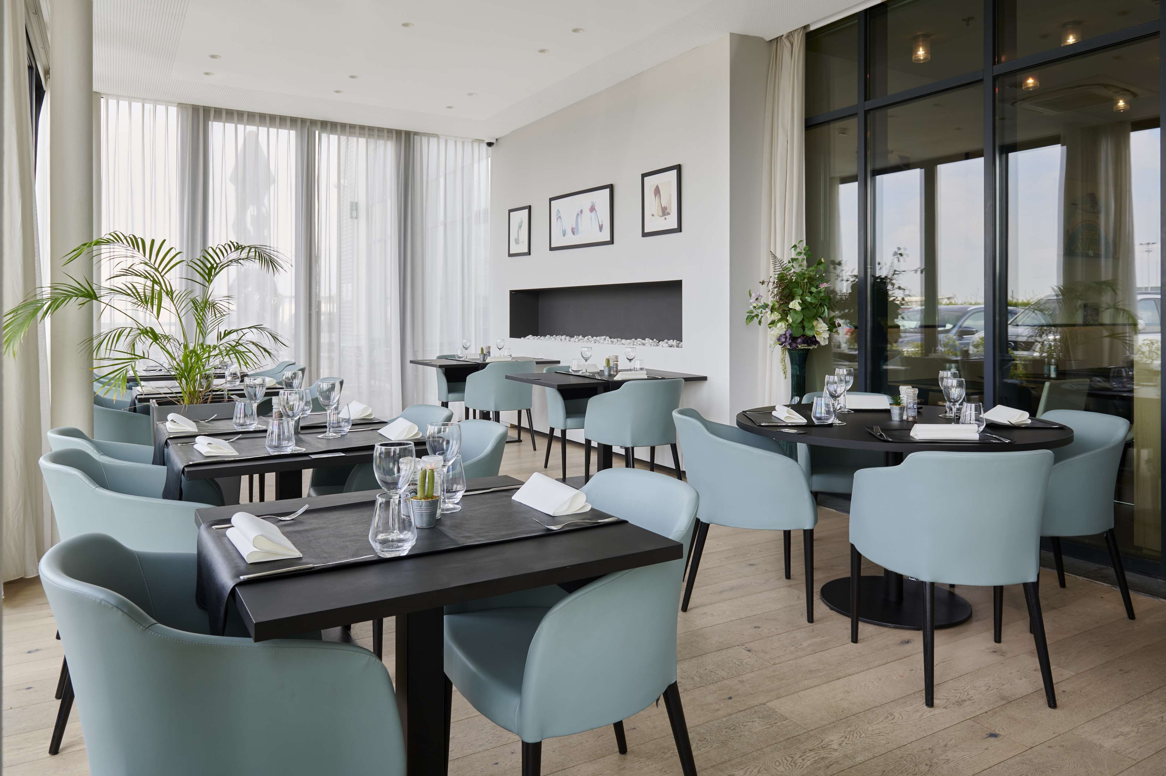 Un espace de restauration moderne doté de plusieurs tables et chaises, d’une cheminée et de grandes fenêtres offrant une vue sur la ville.