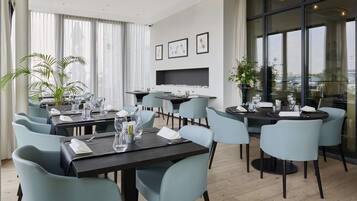 Un espace de restauration moderne doté de plusieurs tables et chaises, d’une cheminée et de grandes fenêtres offrant une vue sur la ville.