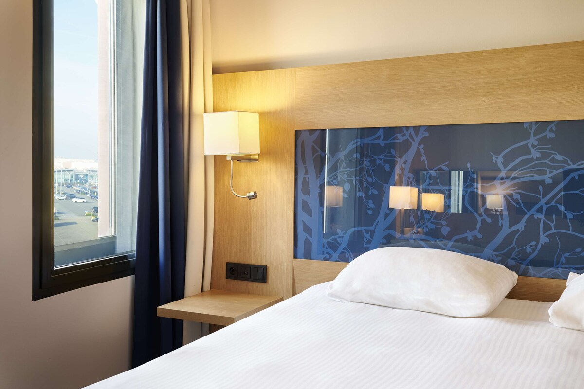 Habitación de hotel con cama, mesita de noche, lámpara y vista a la ciudad a través de una ventana.