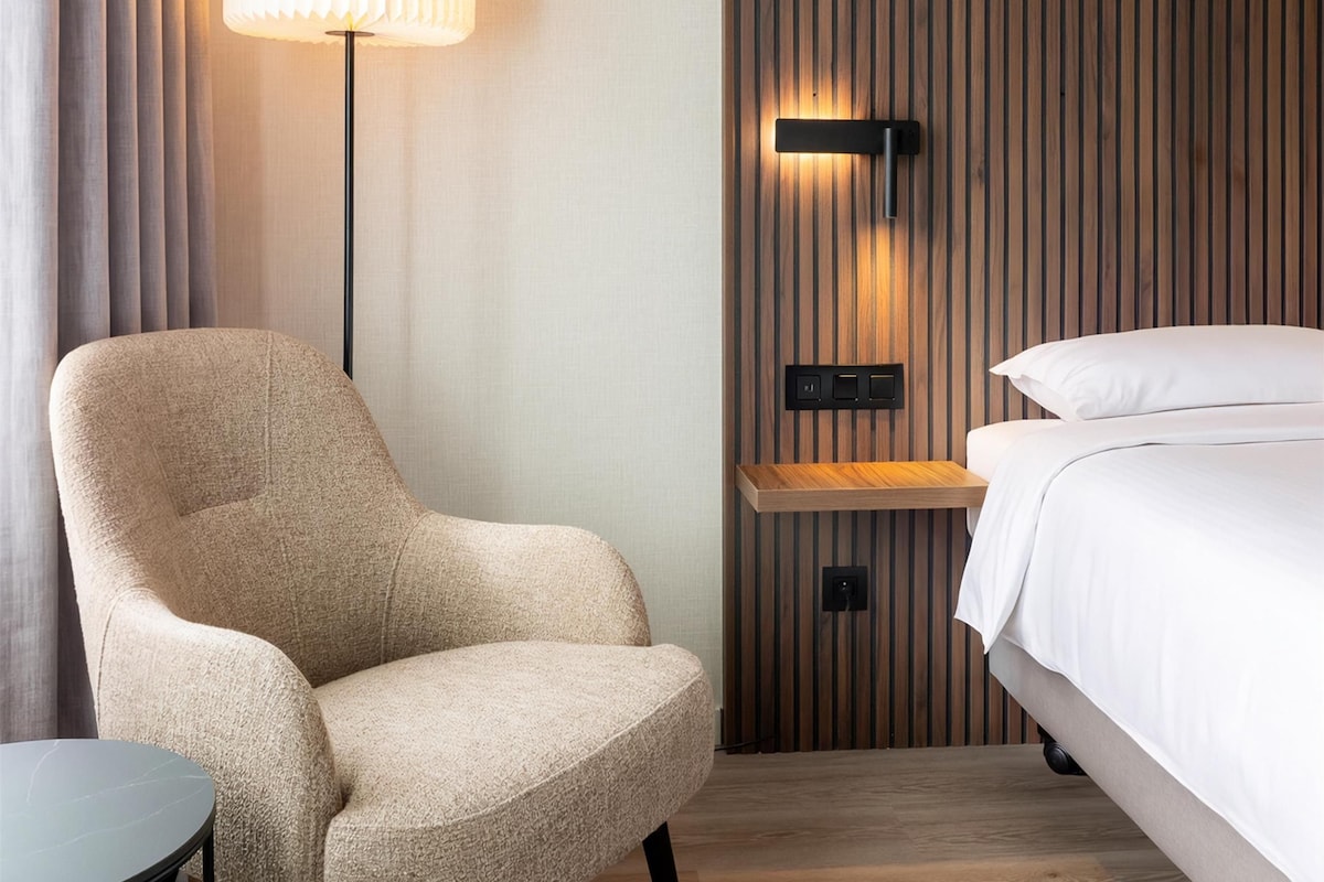 Une chambre d’hôtel comprenant un fauteuil, une table de chevet, une lampe, un lit et une étagère fixée au mur.