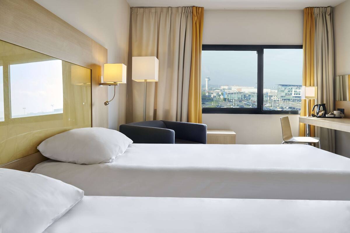 A hotel room with two beds, a desk, a chair, and a view of the city through a large window.