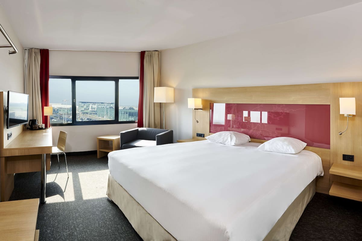 A hotel room with a large bed, a desk with a chair, a TV, and a view of the city.