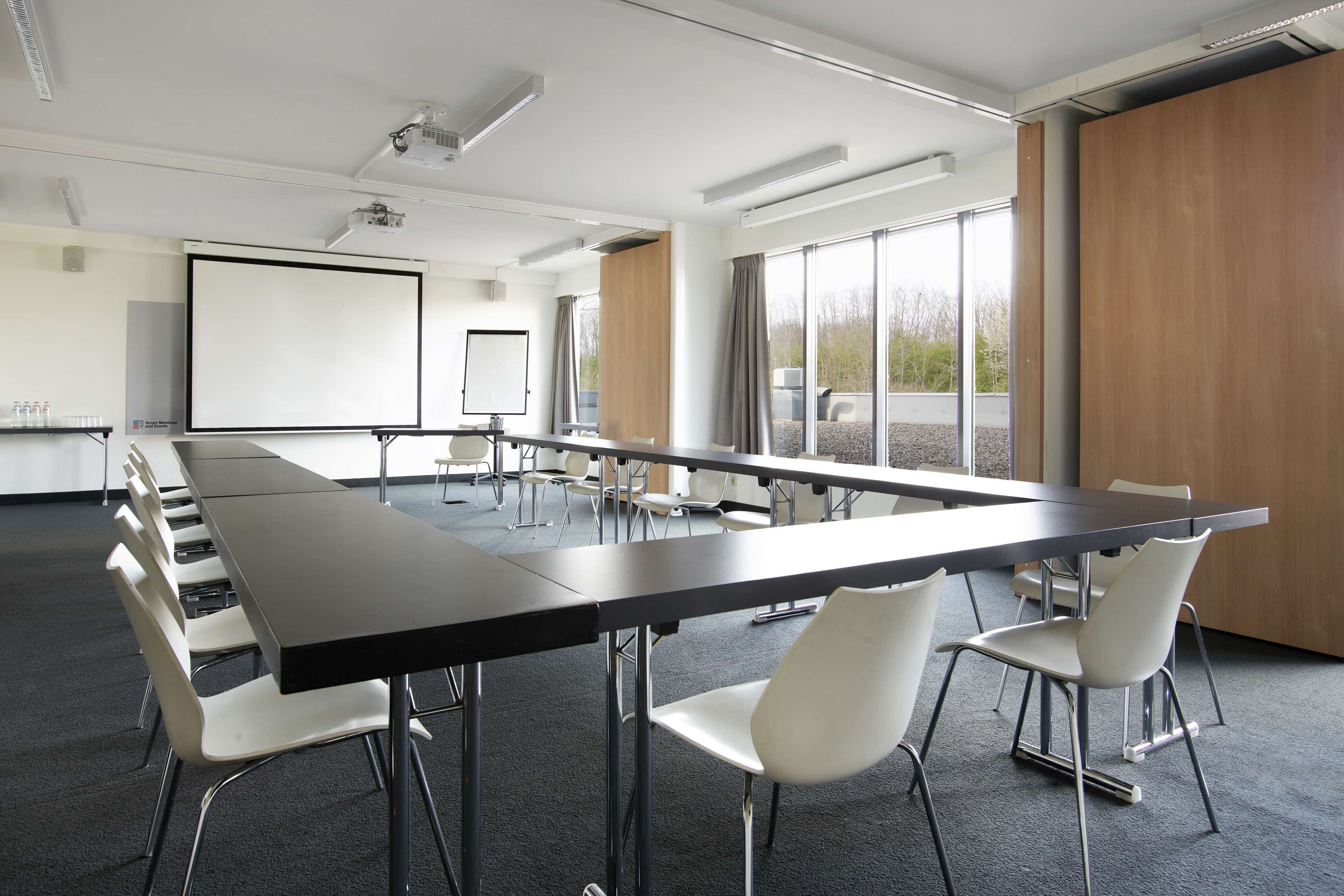 Een vergaderzaal met een lange tafel, witte stoelen, een scherm voor een beamer en grote ramen.