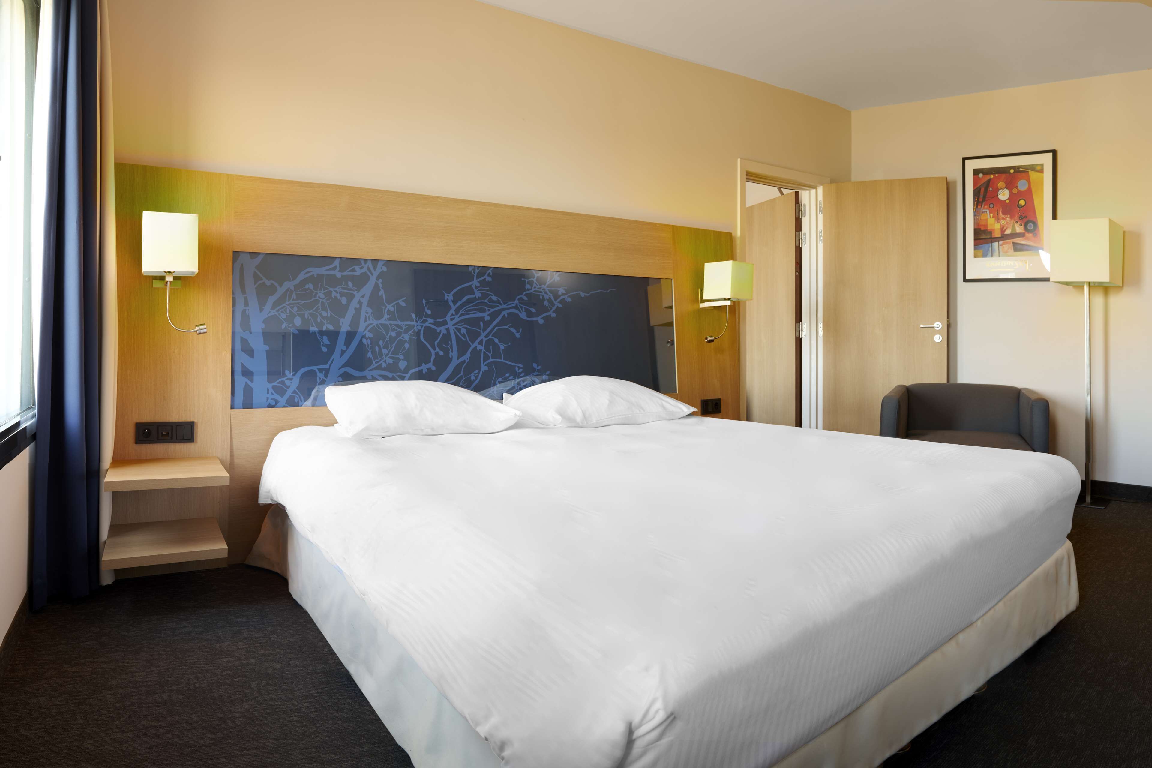 Habitación de hotel con una cama grande, cabecera con diseño de árbol, dos lámparas de noche, una silla y una puerta.