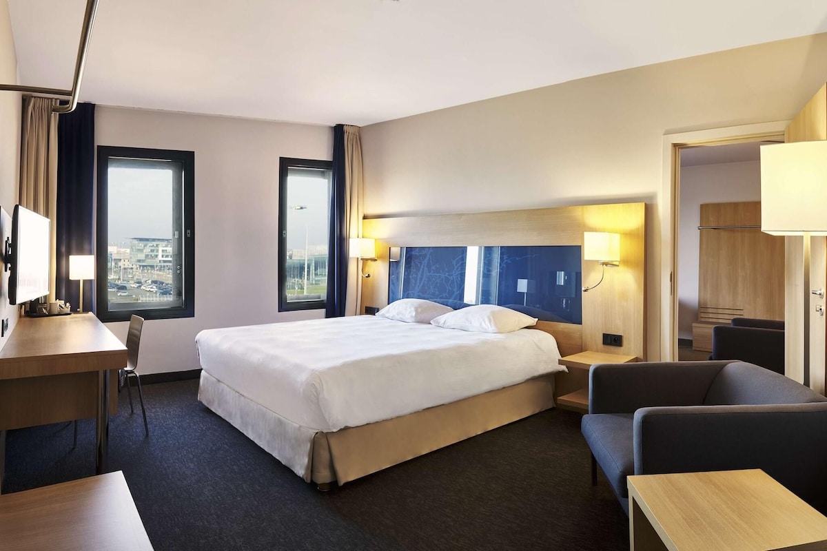 Un cuarto de hotel con una cama grande, un escritorio, una silla y vista a la ciudad por la ventana.