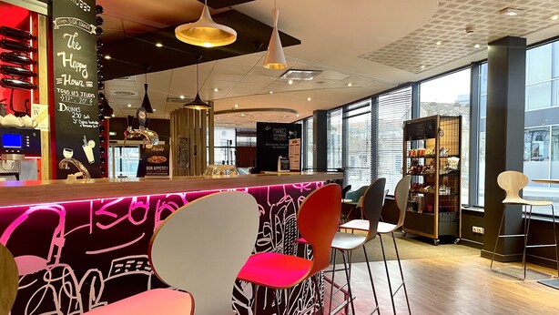 Un espace bar moderne avec des chaises colorées, un comptoir illuminé par des néons et de grandes fenêtres laissant entrer la lumière naturelle.