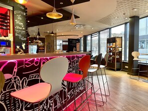 Un área de bar moderna con sillas de colores, una barra iluminada con neón y grandes ventanales que dejan entrar luz natural.
