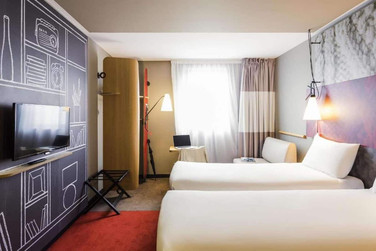 Een hotelkamer met twee bedden, een televisie, een bureau met een laptop en een raam met gordijnen.