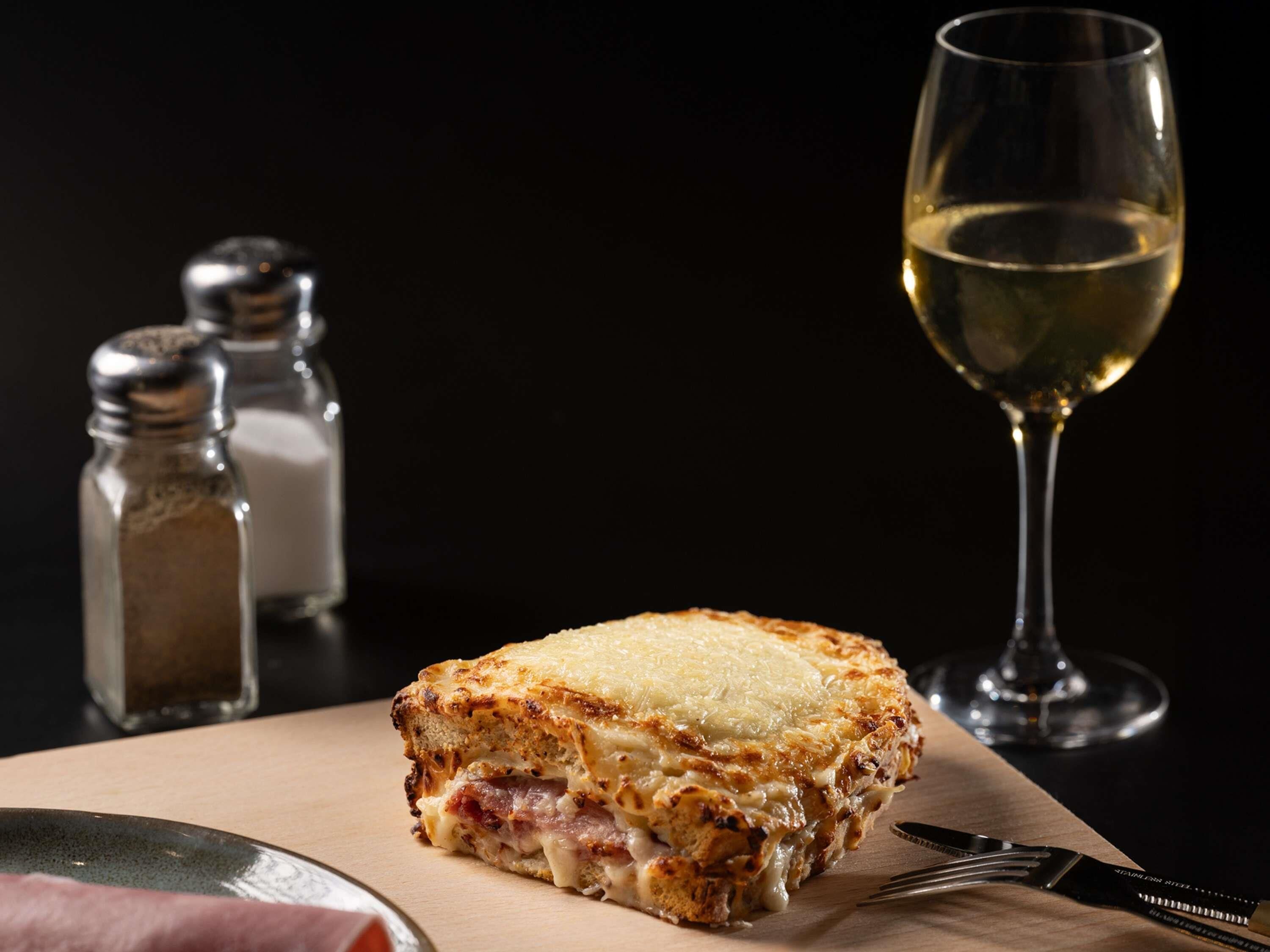 A sandwich with melted cheese and ham, a glass of white wine, and salt and pepper shakers on a wooden board.