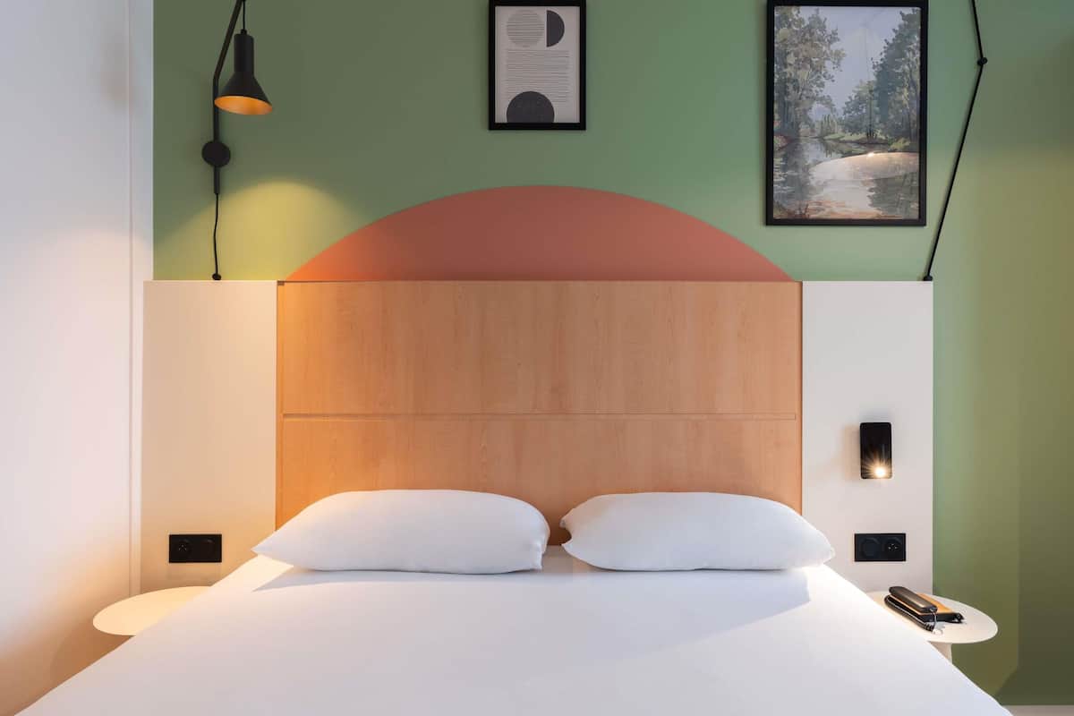 Un dormitorio moderno con cabecera de madera, dos almohadas blancas, una mesita de noche con lámpara y obras de arte enmarcadas en las paredes.