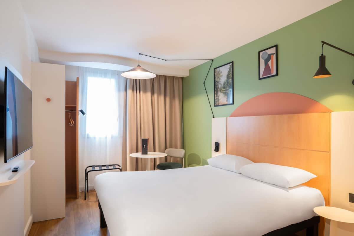 Habitación de hotel con una cama grande, un televisor sobre un mueble bajo, una mesita pequeña y una silla.