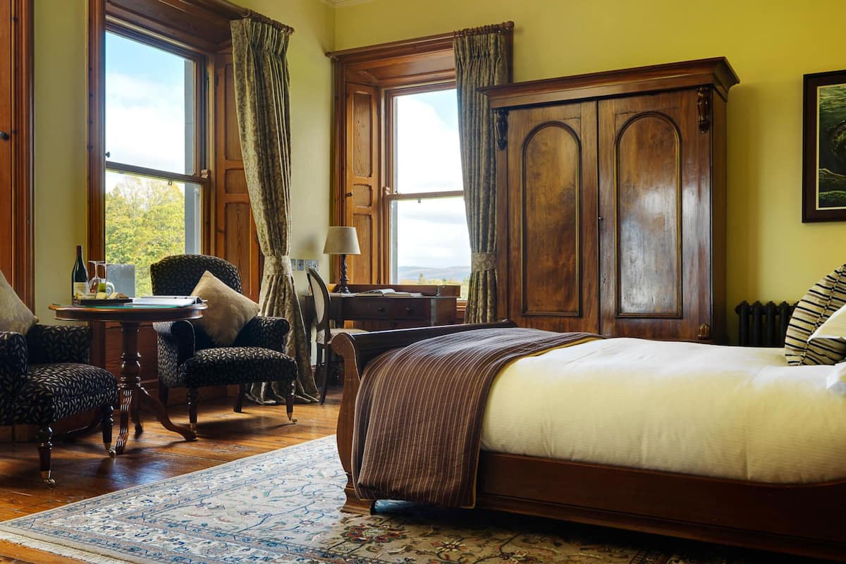 A traditional room with a large bed, a wooden wardrobe, a desk with a lamp, and a view of the outdoors through large windows.