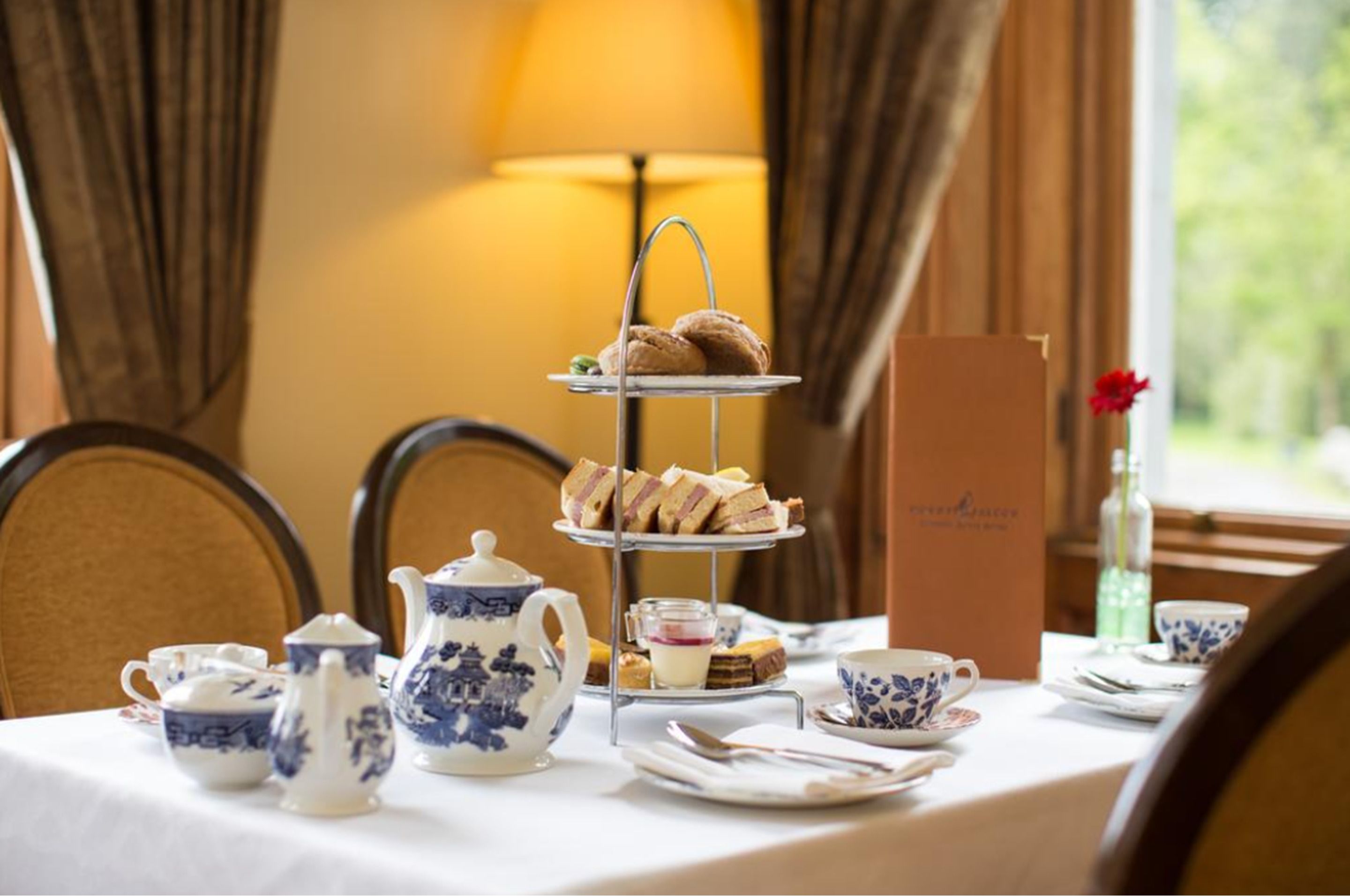A three-tiered dessert stand with pastries, a teapot, cups, and a vase with a flower on a table.