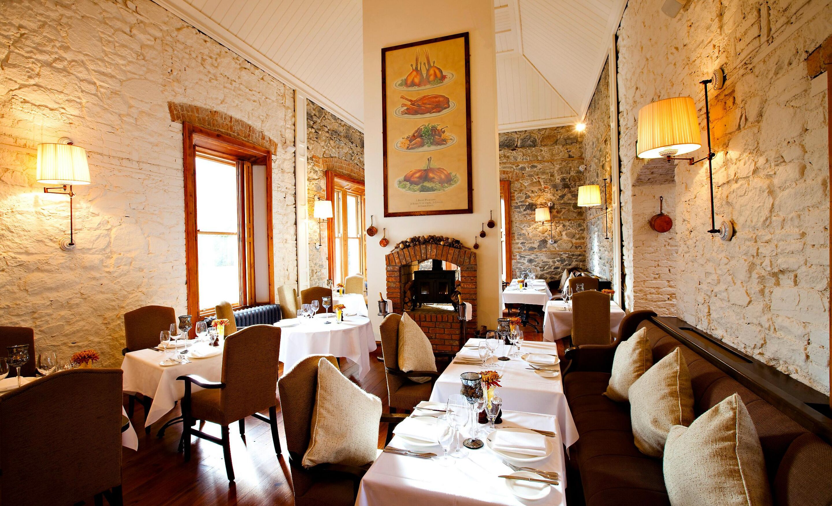 A restaurant with stone walls, wooden beams, and a fireplace. Tables are set with white tablecloths and silverware.