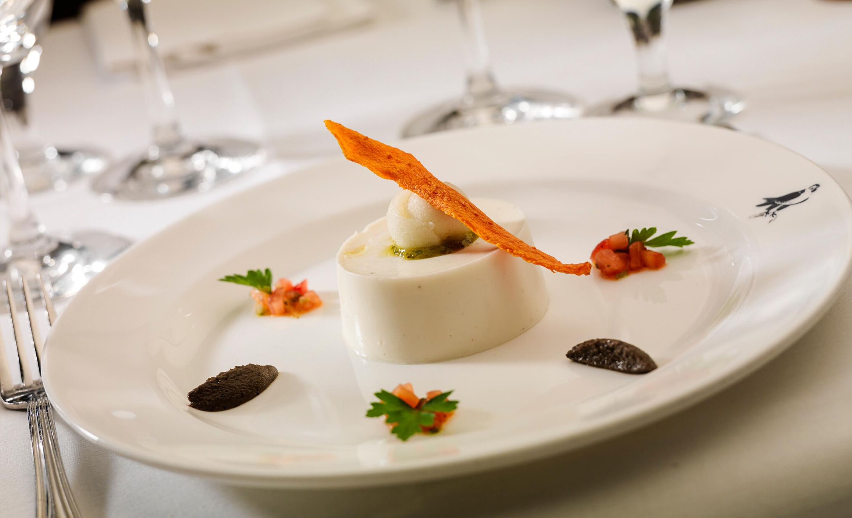 A white plate with a dessert centerpiece, garnished with parsley and a crispy element, surrounded by small garnishes and a dark chocolate piece.