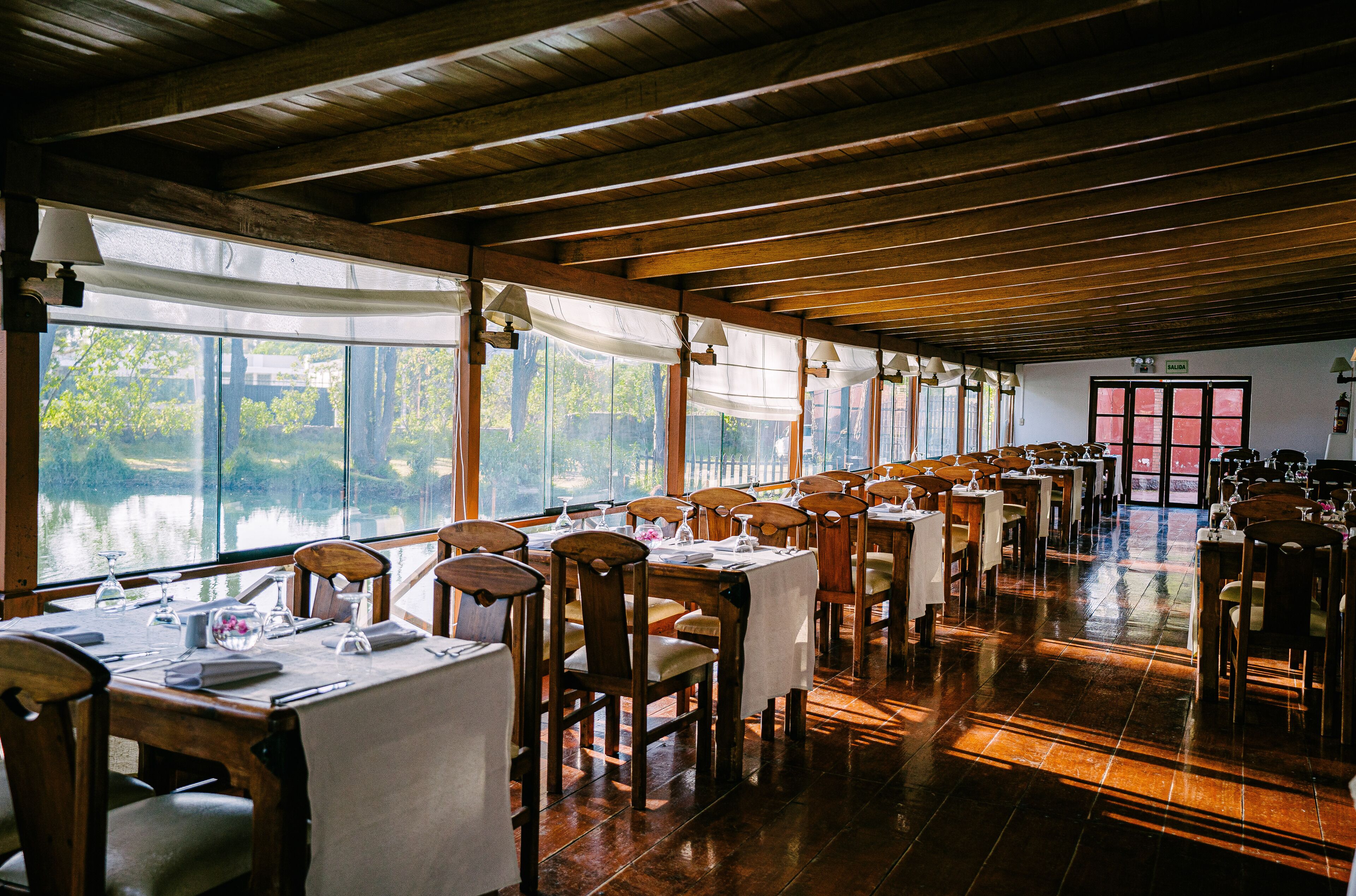 A restaurant with wooden tables and chairs, large windows overlooking a body of water, and a wooden ceiling.