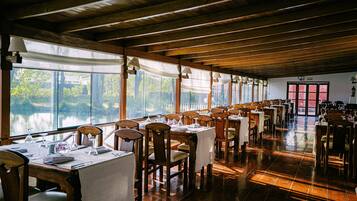 A restaurant with wooden tables and chairs, large windows overlooking a body of water, and a wooden ceiling.