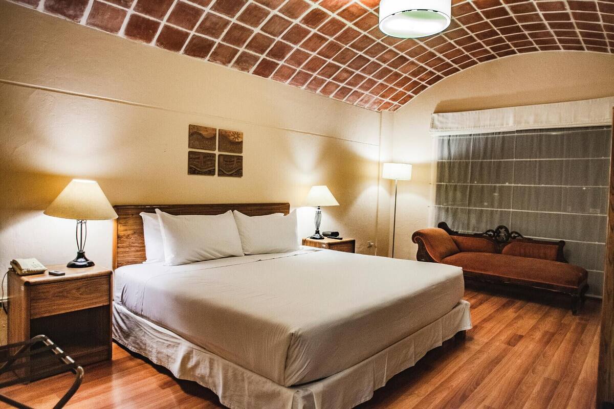 Quarto de hotel com uma cama grande, duas mesas de cabeceira com luminárias, piso de madeira e um sofá.