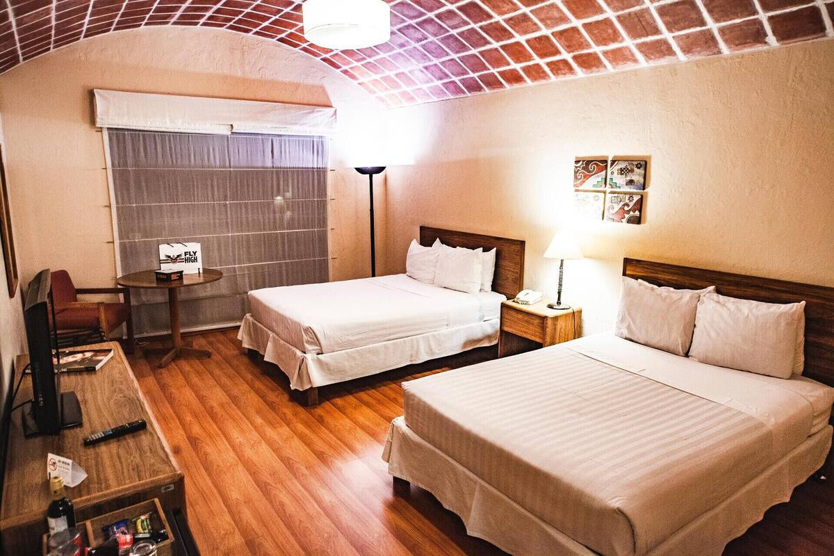 Quarto de hotel com duas camas, uma escrivaninha com cadeira, uma mesinha e um prateleira na parede com quadros.