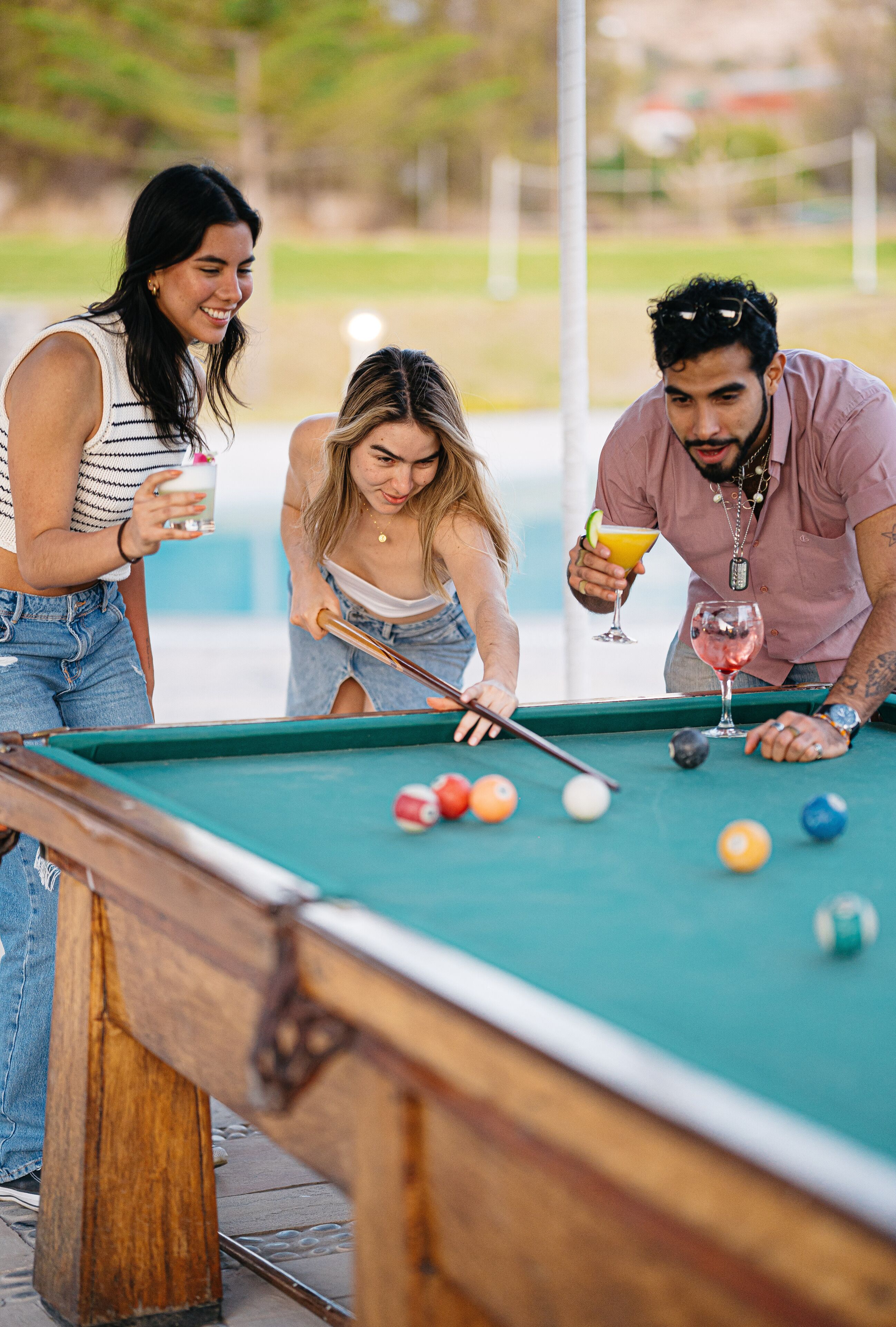 A pool table with three people playing, a green pool table, and a drink in each hand.