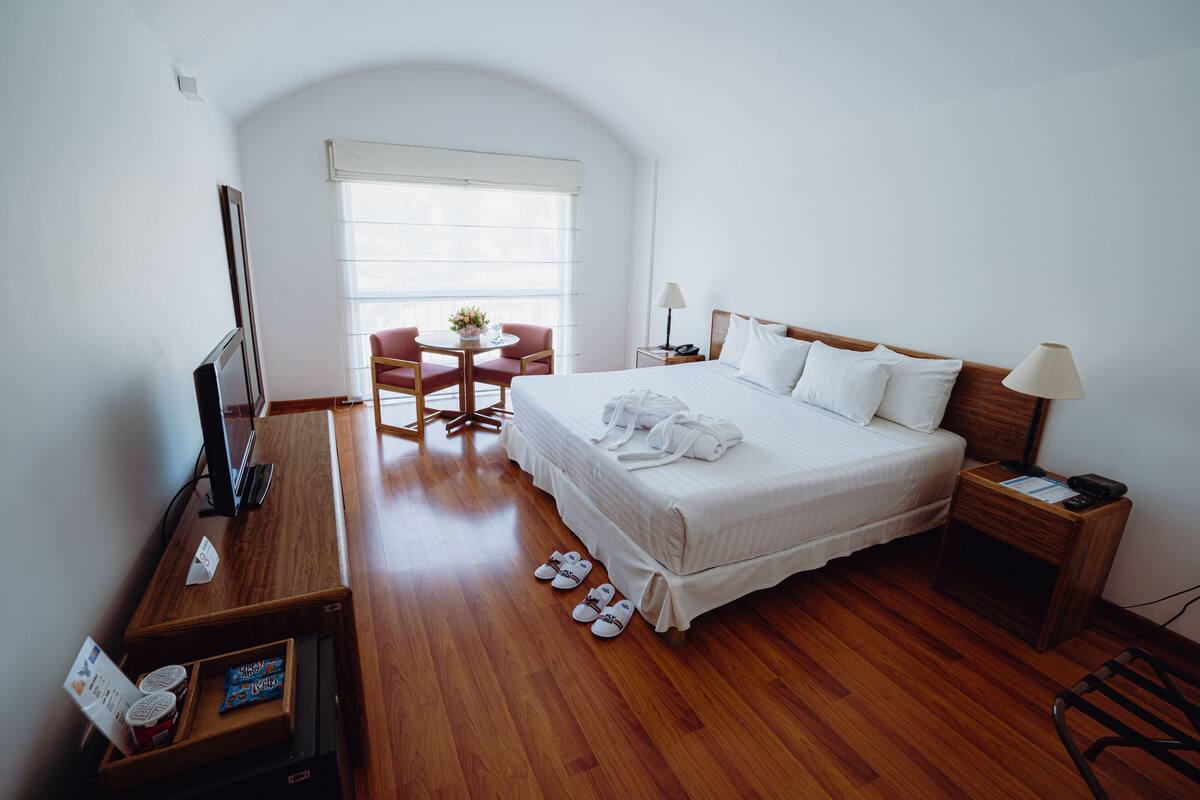 Quarto de hotel com uma cama grande, uma mesa com um computador, uma cadeira, uma mesinha de centro com um vaso e uma janela com persianas.