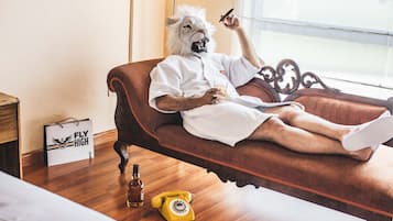 Una persona con máscara de león recostada sobre un sofá marrón tapizado en una habitación con suelo de madera, una ventana y una botella de licor en el piso.