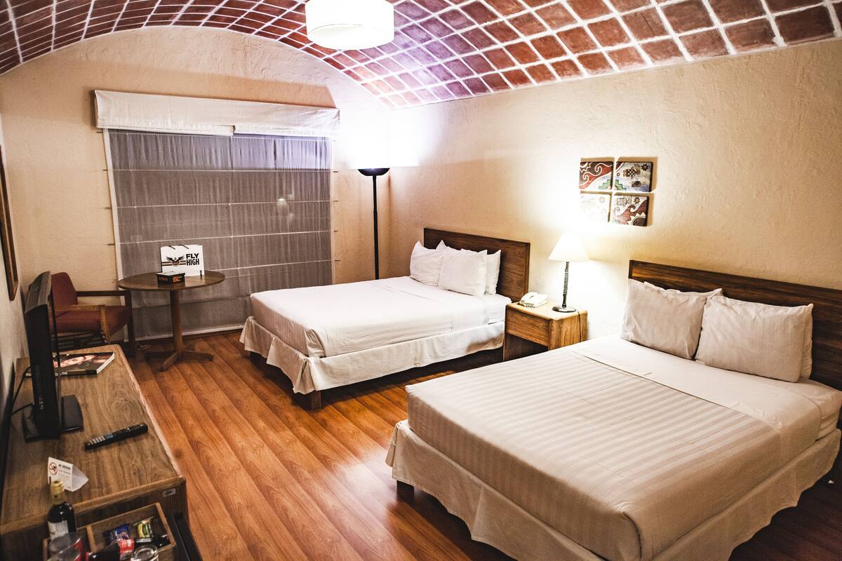 Quarto de hotel com duas camas, uma escrivaninha, uma cadeira, um abajur e uma mesinha de cabeceira com uma garrafa e um controle remoto.