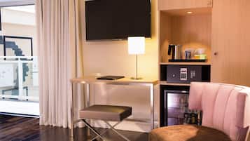 Une chambre d’hôtel compacte comprenant un bureau, un téléviseur à écran plat, un petit réfrigérateur, une machine à café et un coin salon avec un banc rembourré.