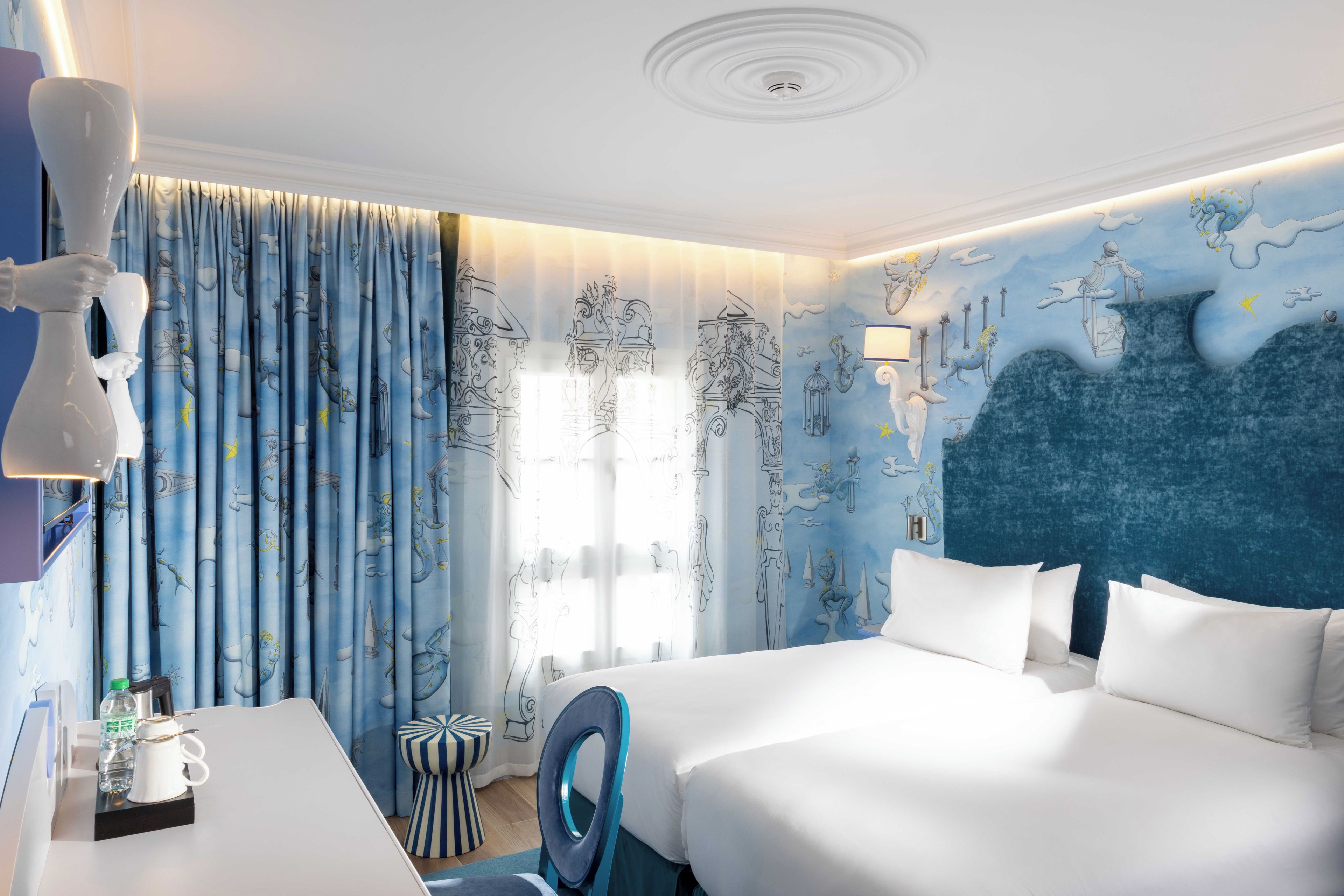 Ett hotellrum med en stor säng, ett skrivbord och en stol. Det finns ett fönster med gardiner, en vägg med en väggmålning och ett tak med en rund lampa.