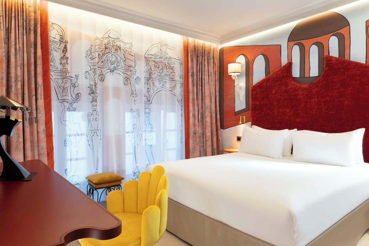 Ein Hotelzimmer mit einem großen Bett, einem Schreibtisch mit Lampe, einem gelben Sessel und einem Fenster mit Vorhängen.
