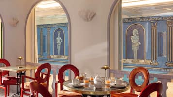 A dining area with round tables, red chairs, and a decorative wall with classical motifs.