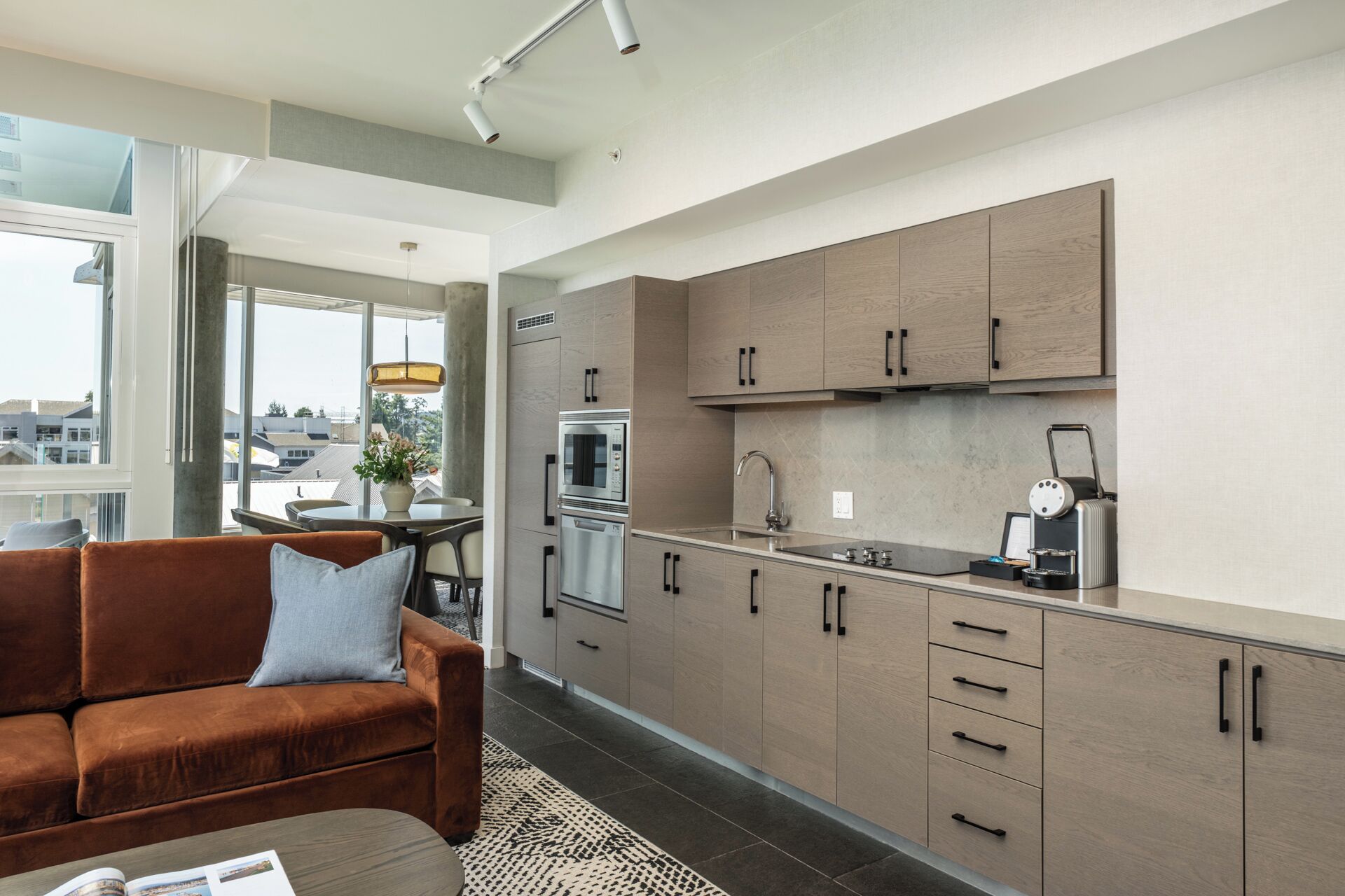 A modern kitchen with built-in appliances, a dining area with a brown sofa, and large windows offering a view of the city.