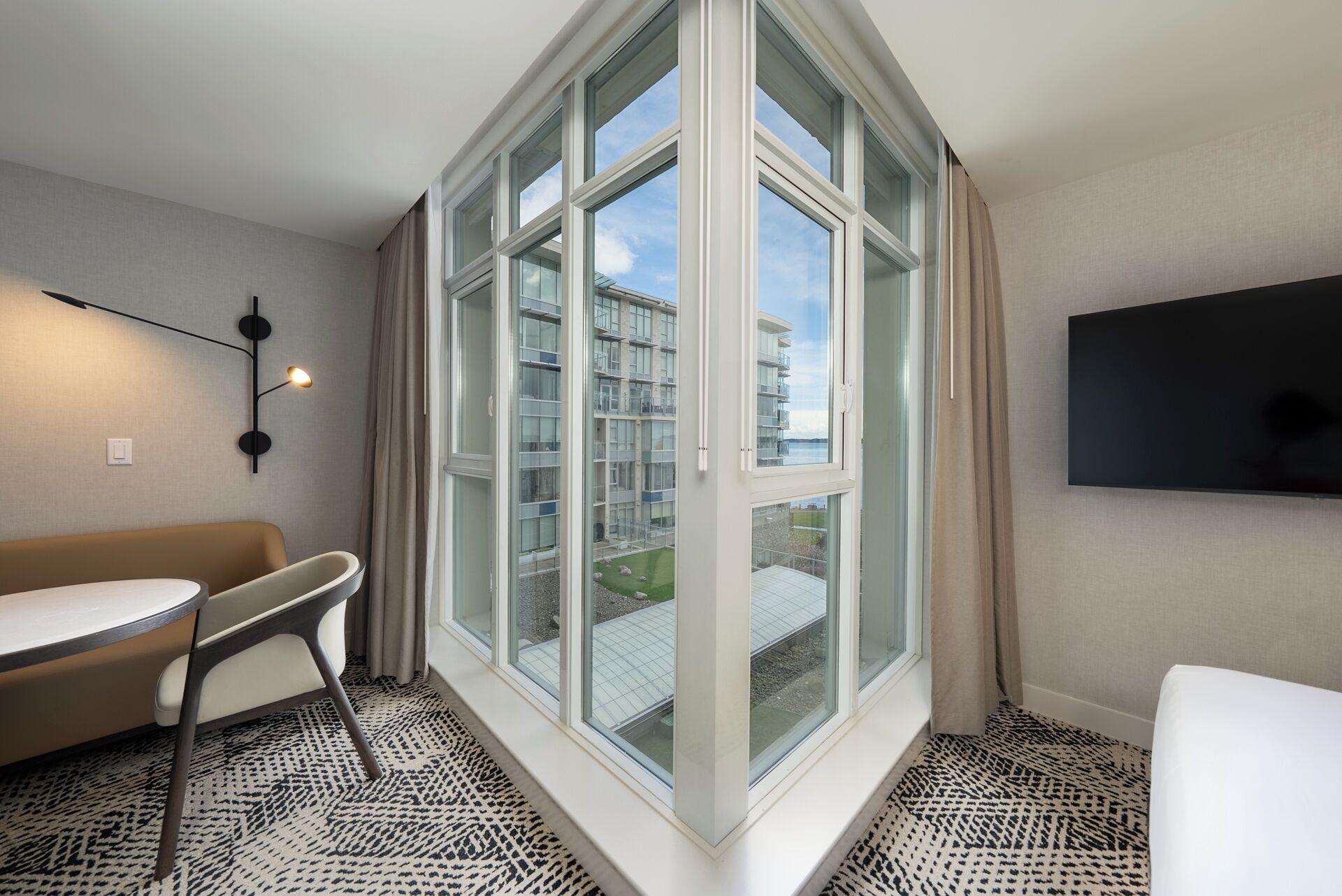Une chambre d’hôtel avec une grande fenêtre, un canapé, une petite table, un téléviseur et une moquette à motifs.