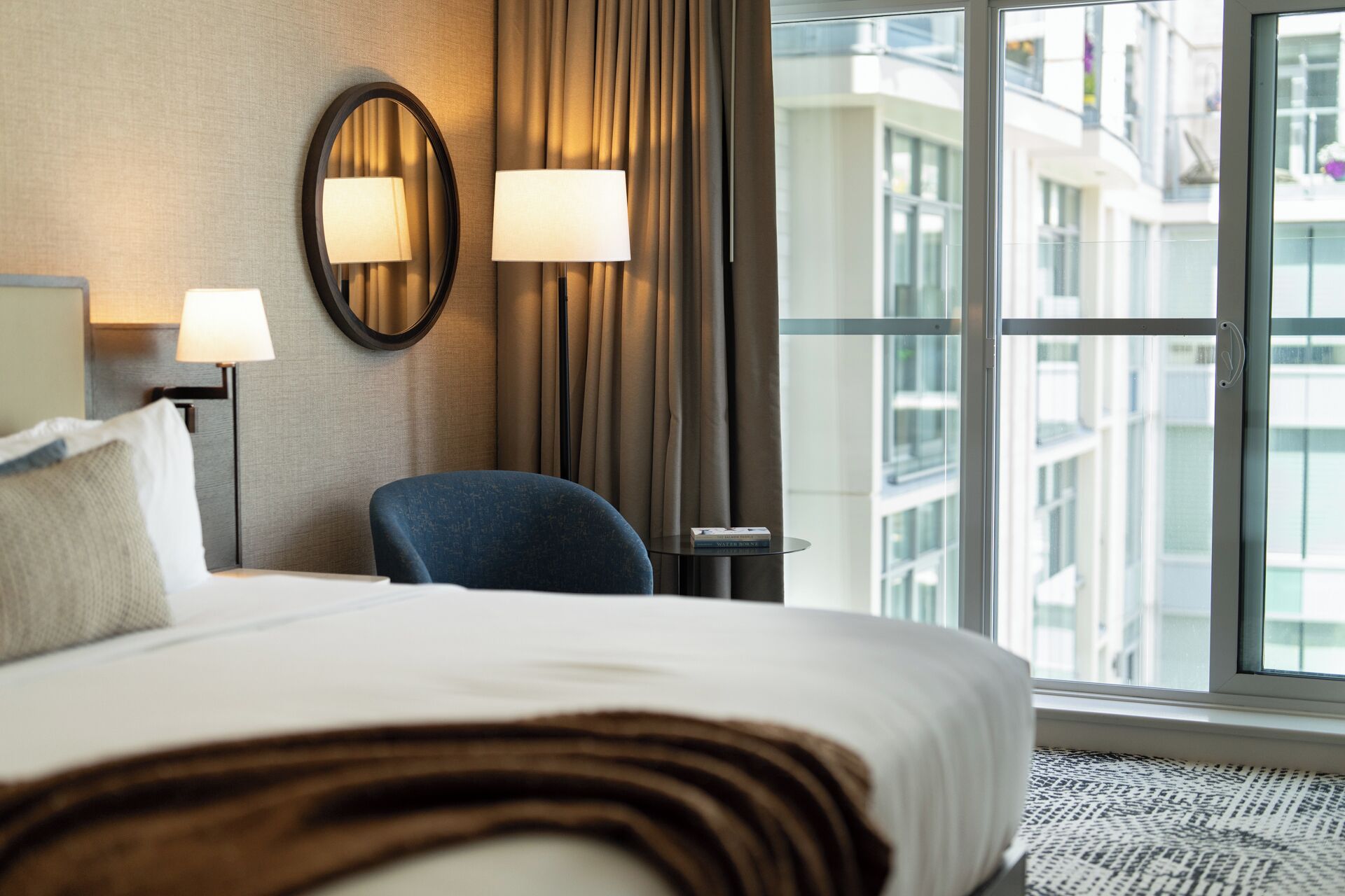 A hotel room with a bed, two wall-mounted lamps, a chair, a small table, and a large window with a view of buildings.