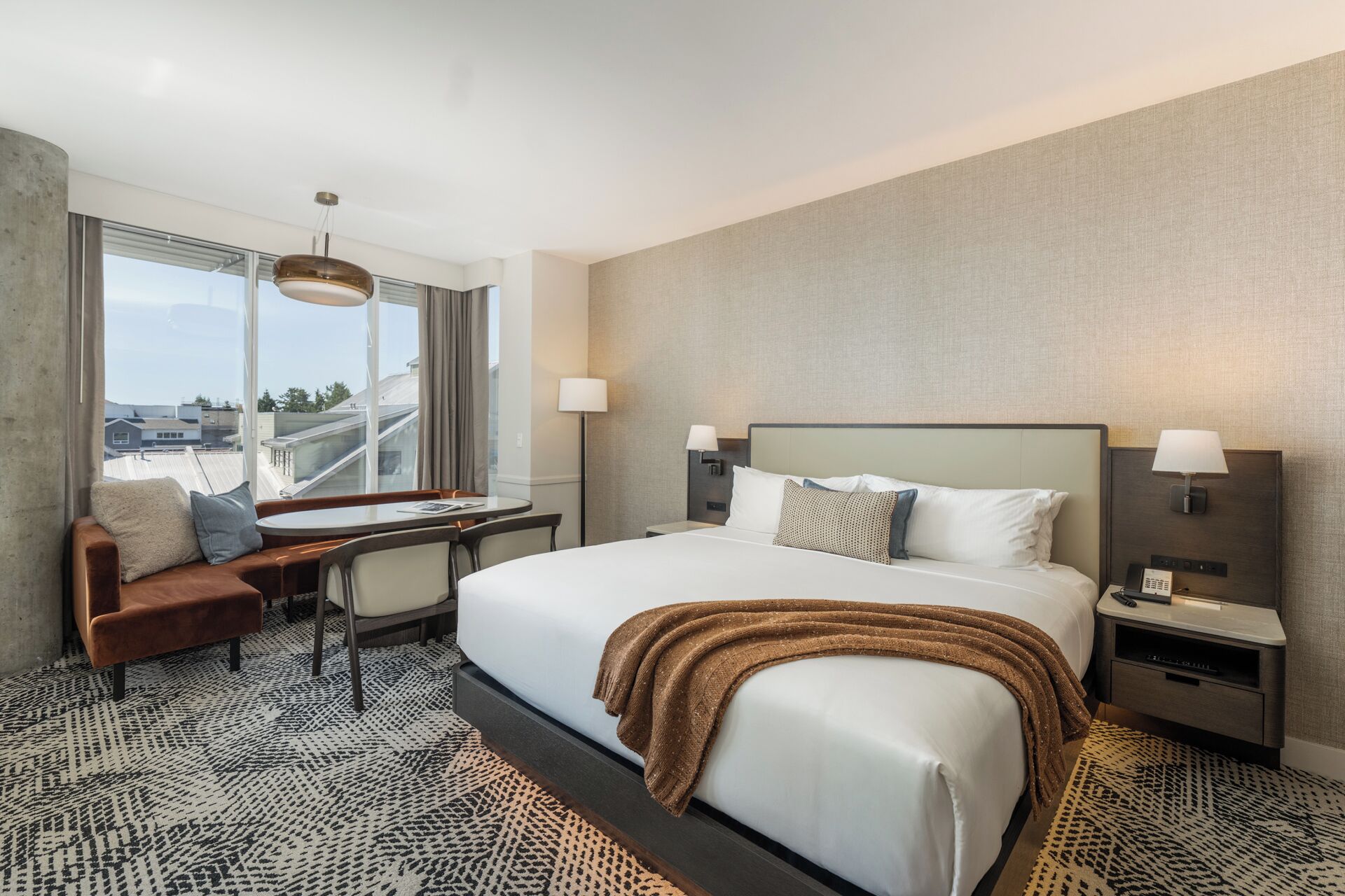 A hotel room with a large bed, two bedside tables, a desk with a chair, a sofa, and a large window offering a view of the cityscape.