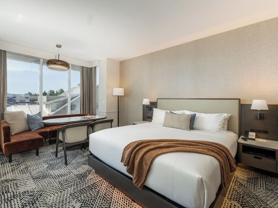 Habitación de hotel con una cama grande, dos mesitas de noche, un escritorio con silla, un sofá y un ventanal con vistas a la ciudad.