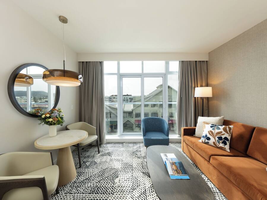 Een hotelkamer met een ronde spiegel, een rond tafel, een blauwe fauteuil, een bruine bank en een kamerkleed met een patroon.