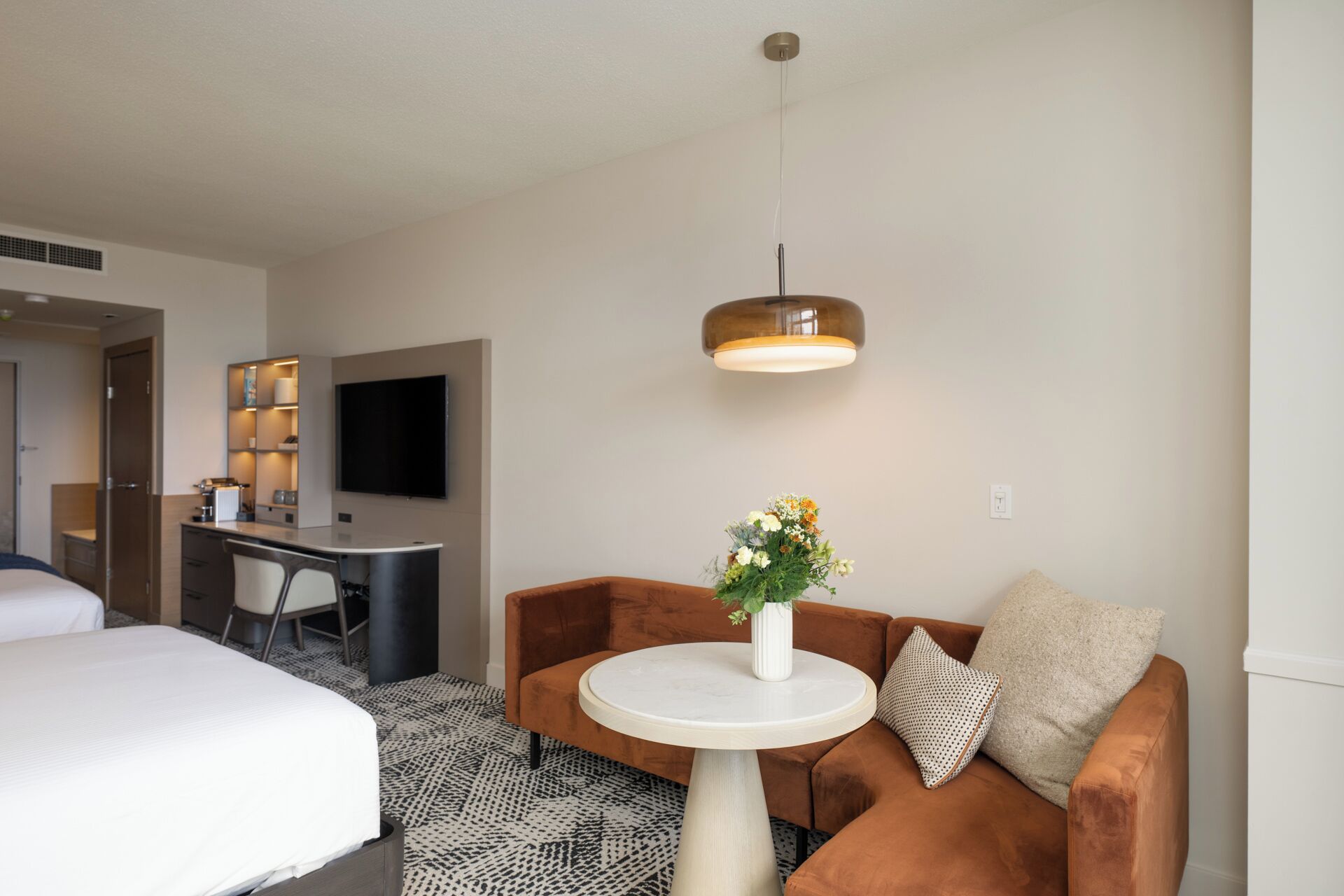 A hotel room with a bed, a sofa, a small round table with a vase of flowers, a desk with a flat-screen TV, and a pendant light.